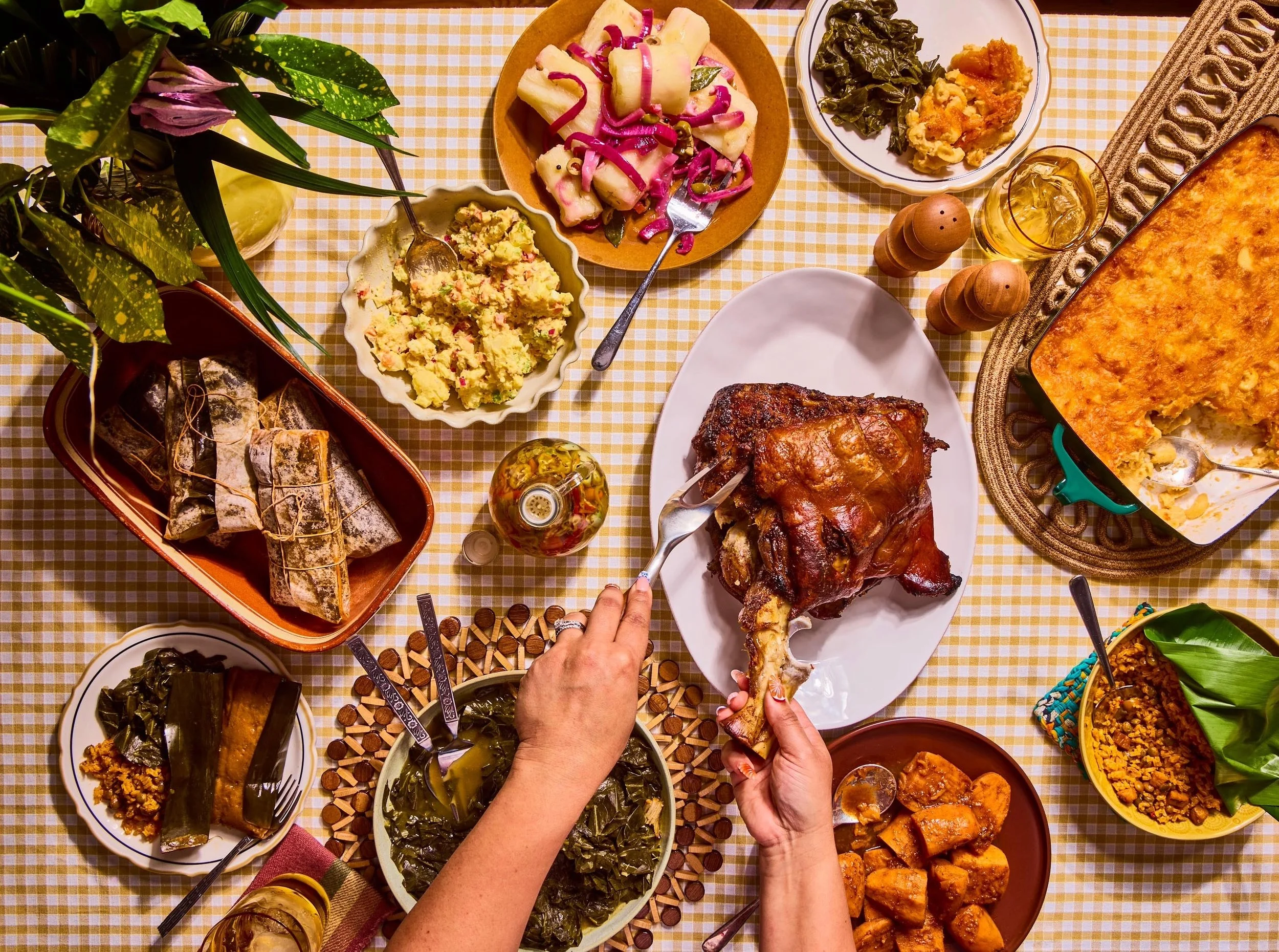 A top-down view of a table with various dishes including mashed potatoes, green vegetables, a casserole, and a glazed ham. There are hands holding a serving fork and a piece of bread or roll, serving food onto a white plate. The tablecloth is yellow 