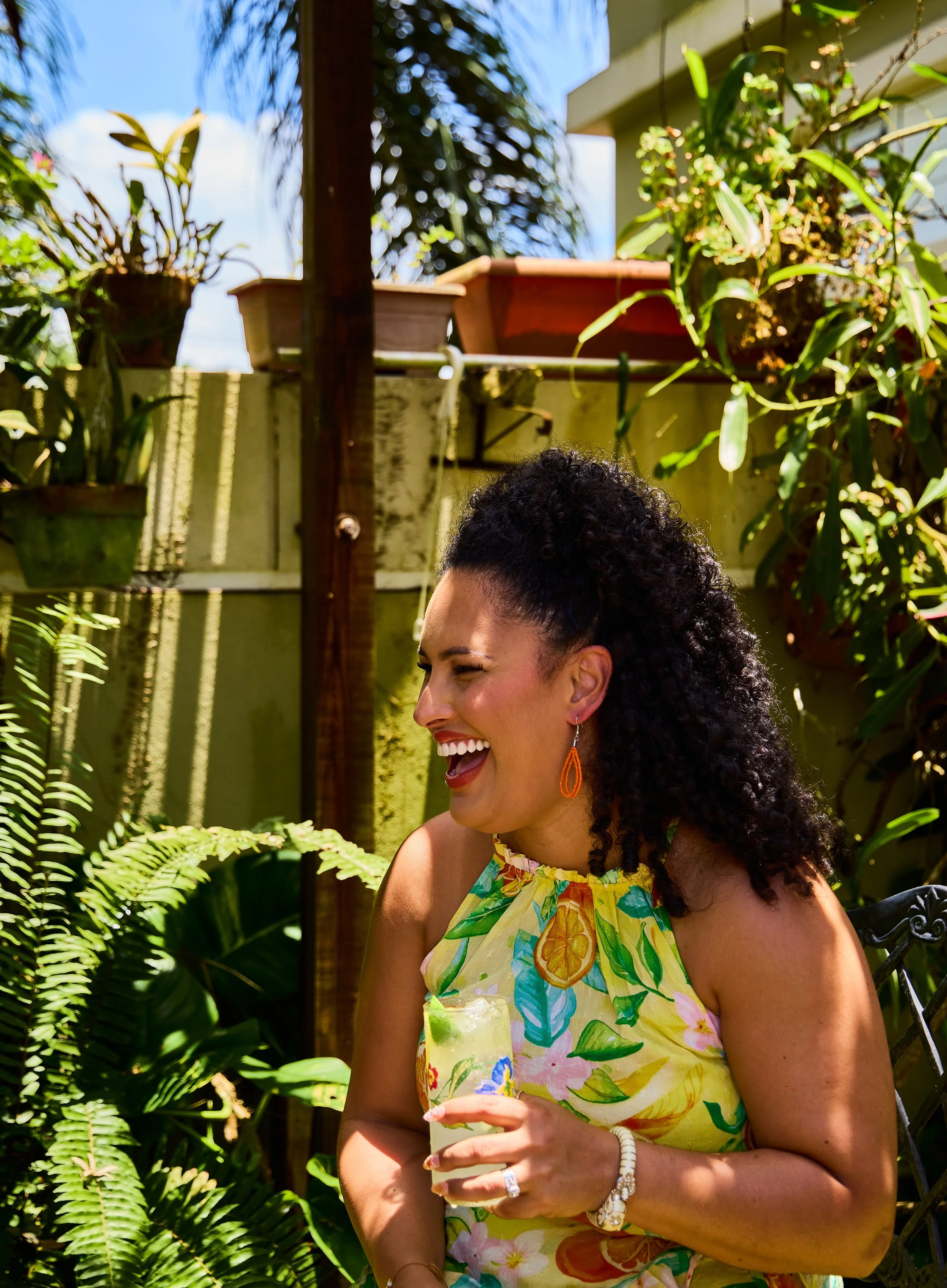 A woman with curly hair smiling and holding a cocktail glass outdoors surrounded by green plants and sunlight.