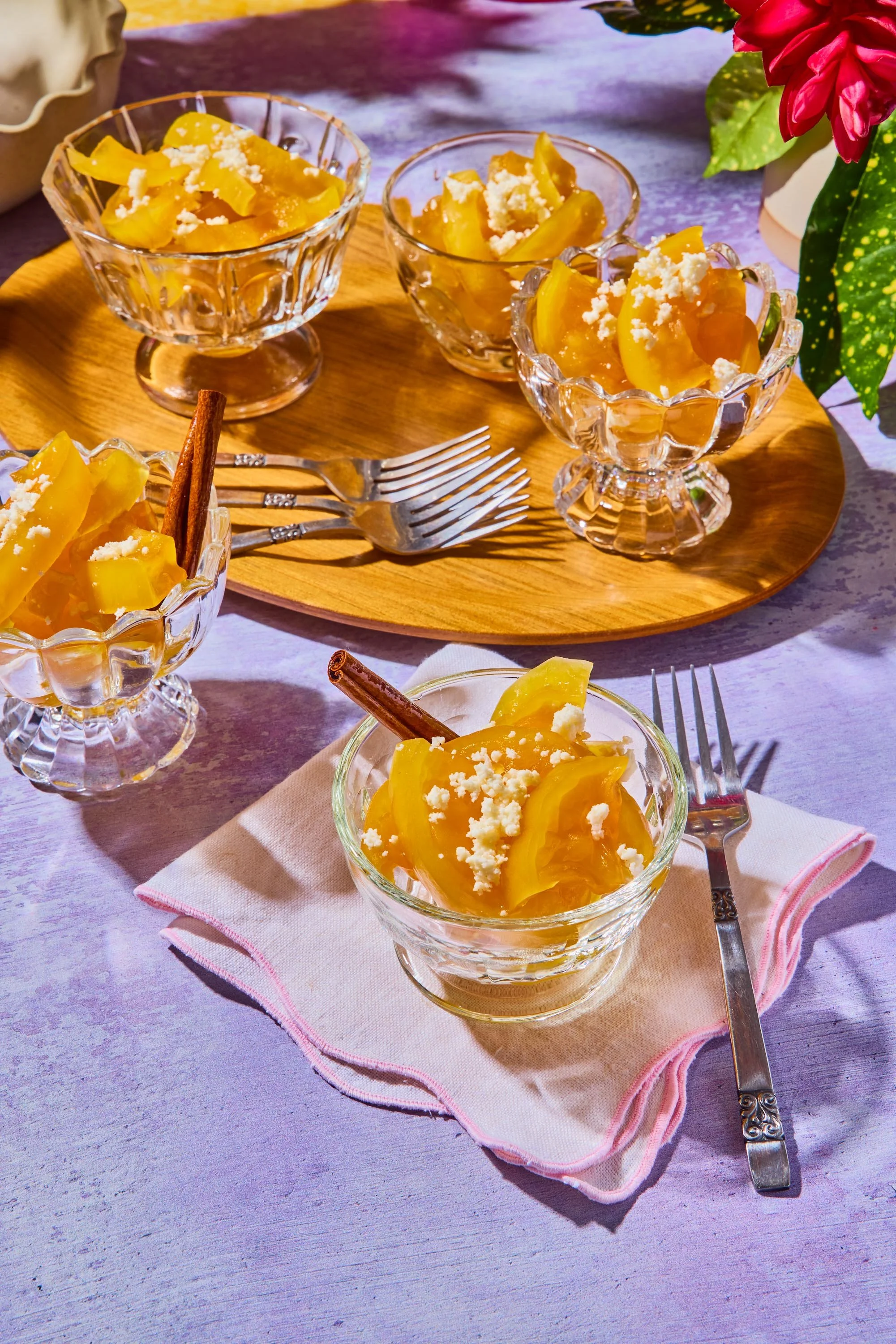 Glass dessert cups filled with yellow jelly or fruit compote, topped with crumbled cheese or white topping, placed on a round wooden tray with forks, set on a pastel pink cloth napkin on a table.