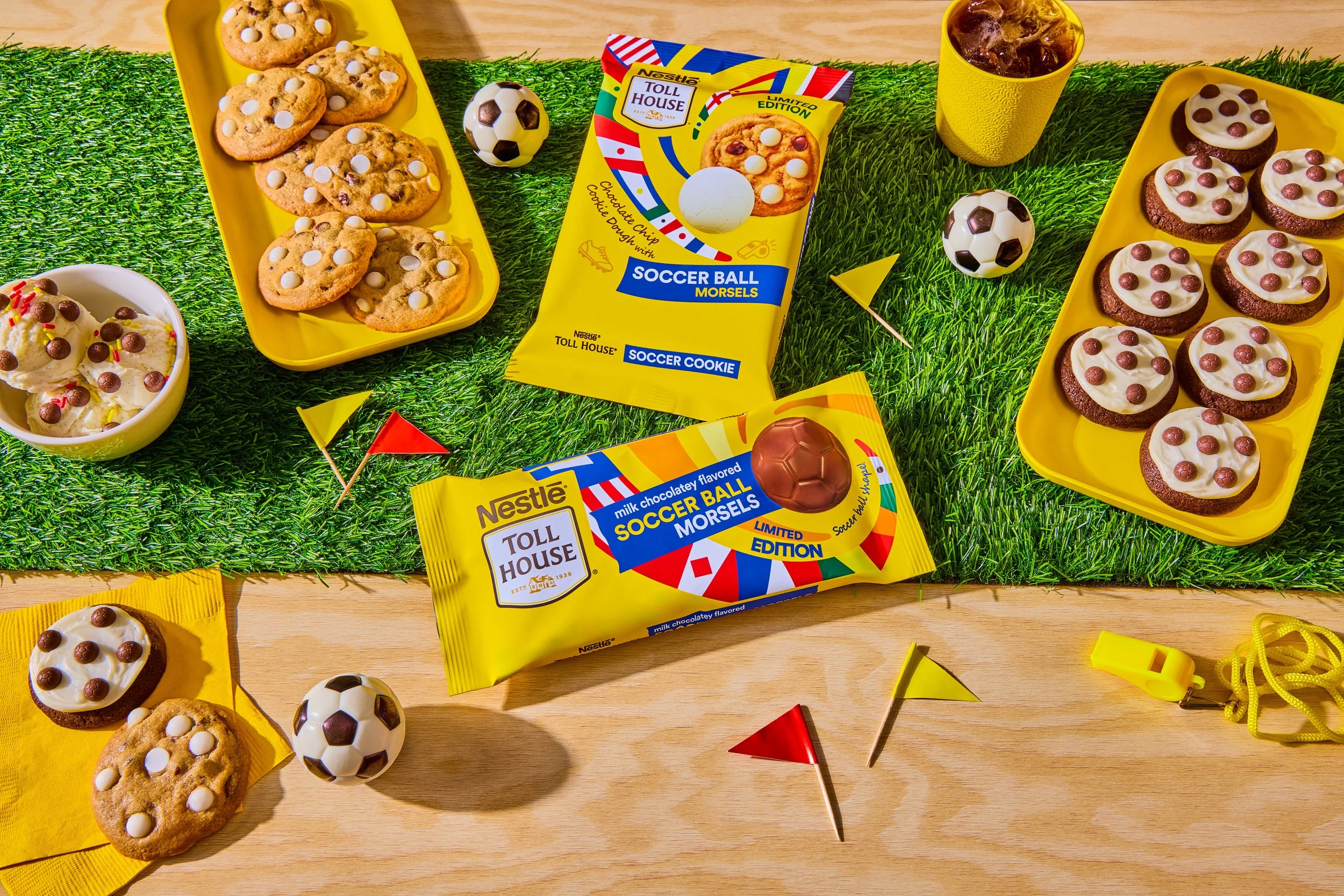 Assorted cookies and chocolate snacks arranged on yellow trays, soccer ball-shaped candies, and small yellow and red flags on a grass and wooden surface.