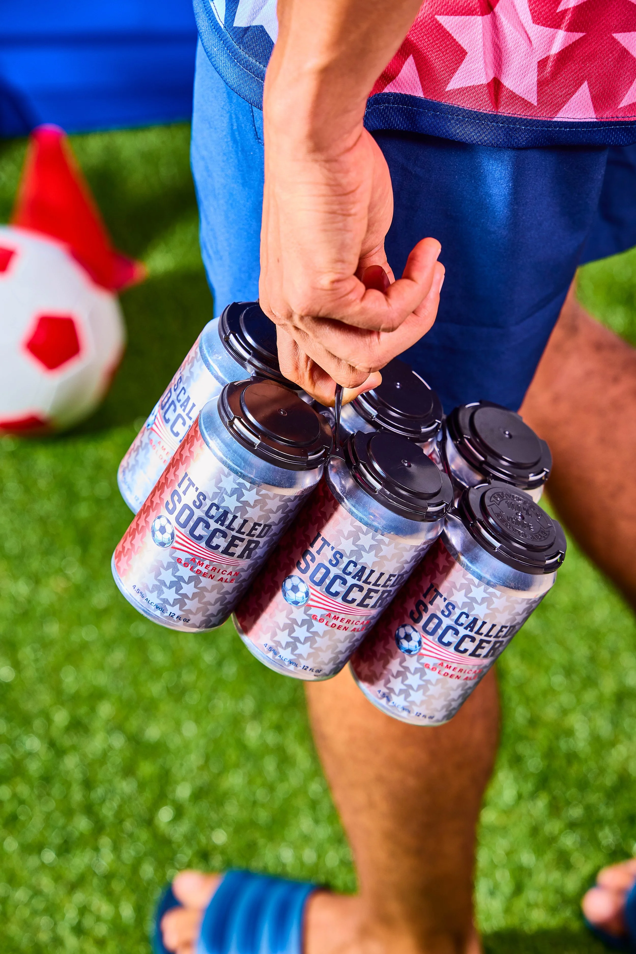 Person holding a six-pack of cans labeled 'It's Called Soccer,' with a soccer ball and a red cone on a grassy field in the background.