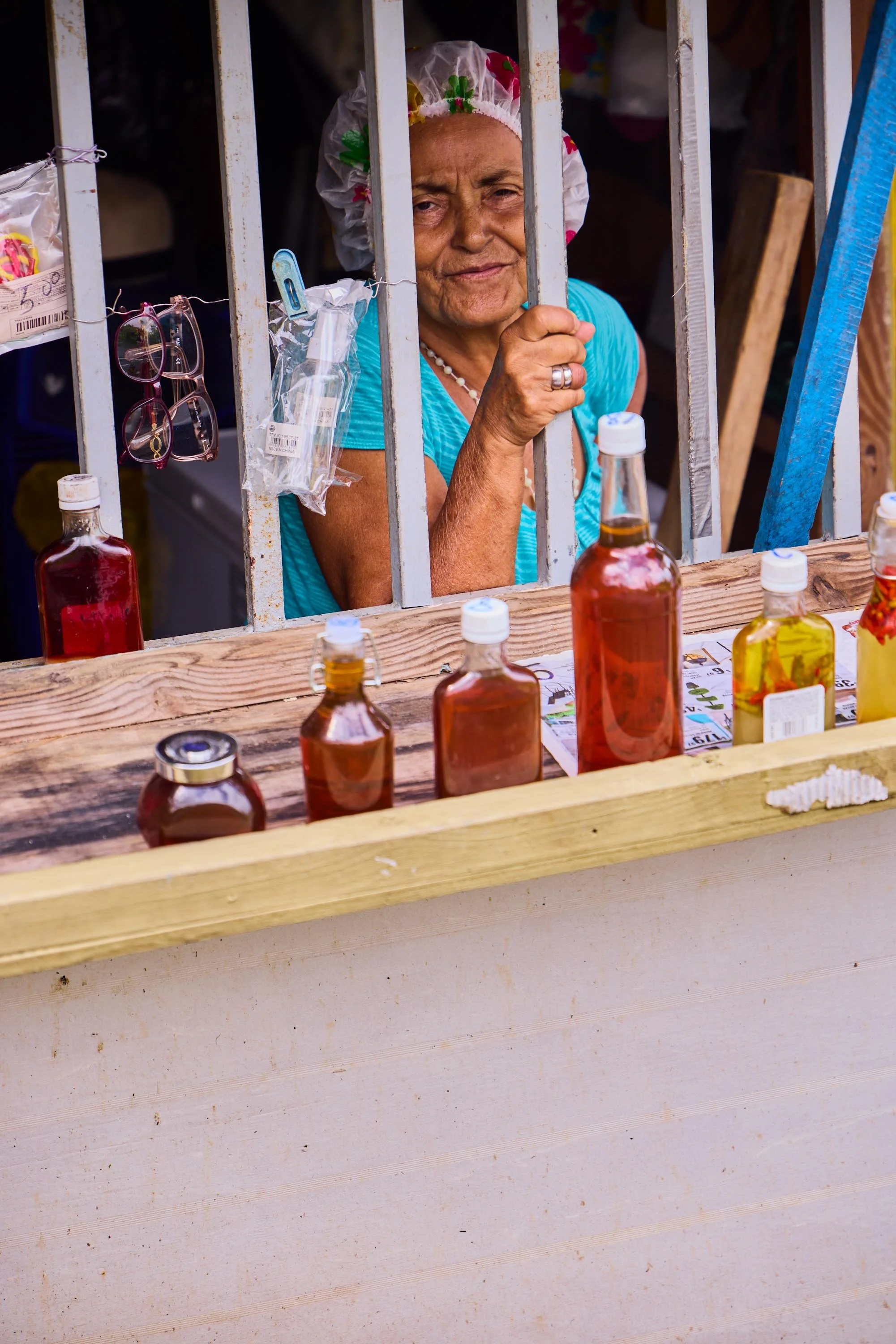 A woman wearing a shower cap is behind metal bars at a small market stall with various bottles of colorful liquids on the wooden counter.