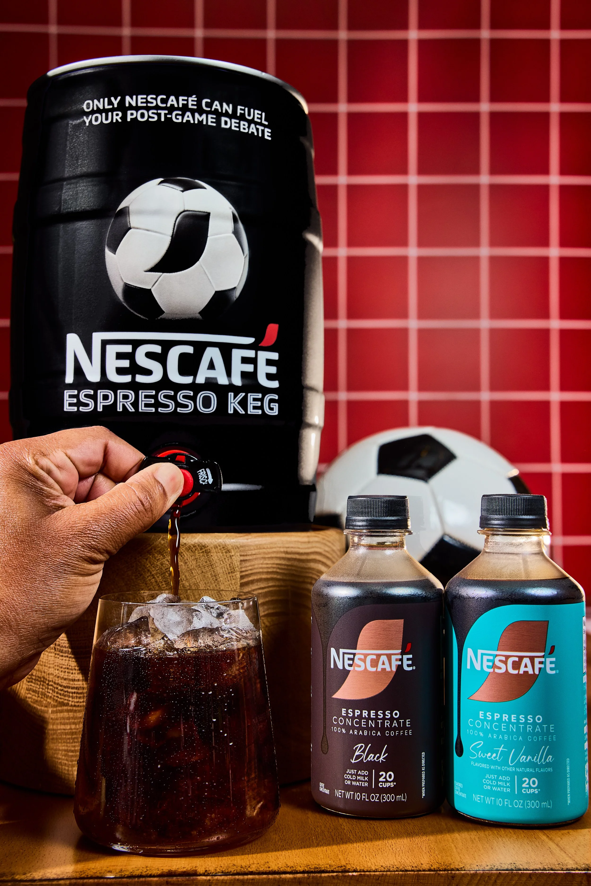 A person is pouring Nescafe espresso into a glass of ice. Behind them are two cans of Nescafe espresso concentrate, one black and one blue, on a wooden surface. There is a soccer ball and a red grid background.