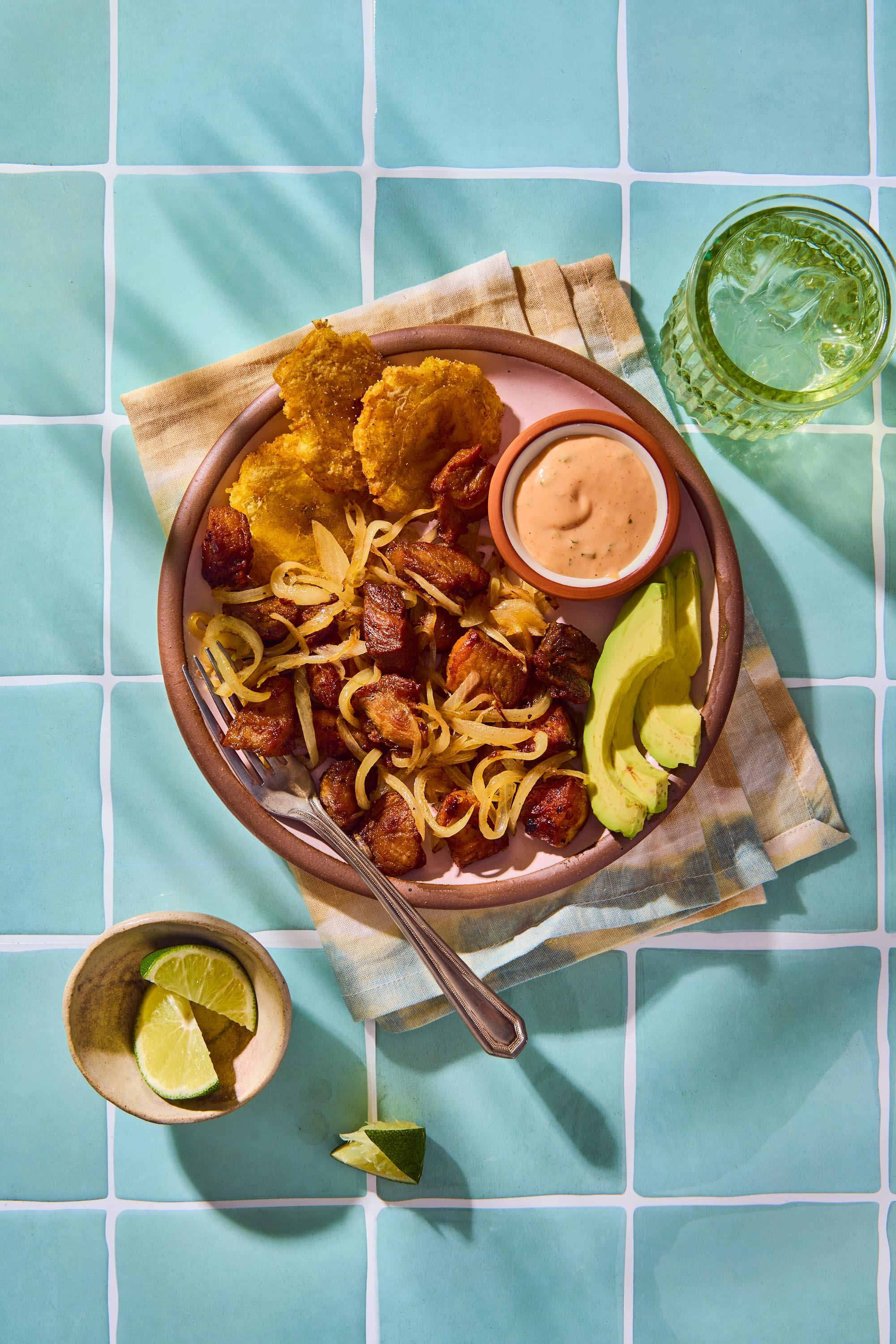 A bowl of Mexican food with fried fish, pork, onions, and a side of avocado slices and a small container of sauce on a light blue tiled table, with lemon wedges on a small dish.