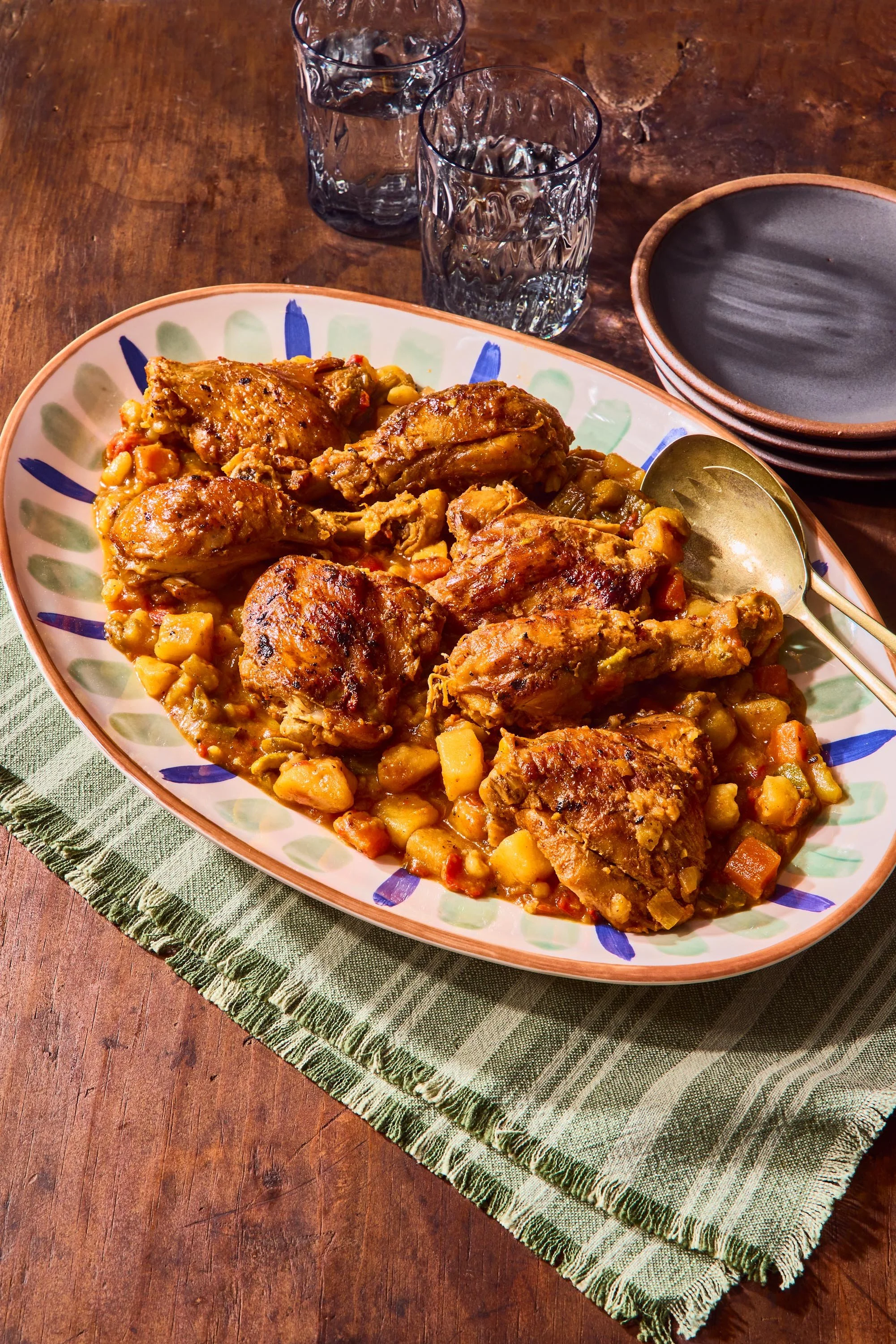 A plate of cooked chicken with potatoes and vegetables on a colorful patterned ceramic dish, set on a green cloth on a wooden table. In the background, there are three glasses of water and a stack of empty dark bowls.