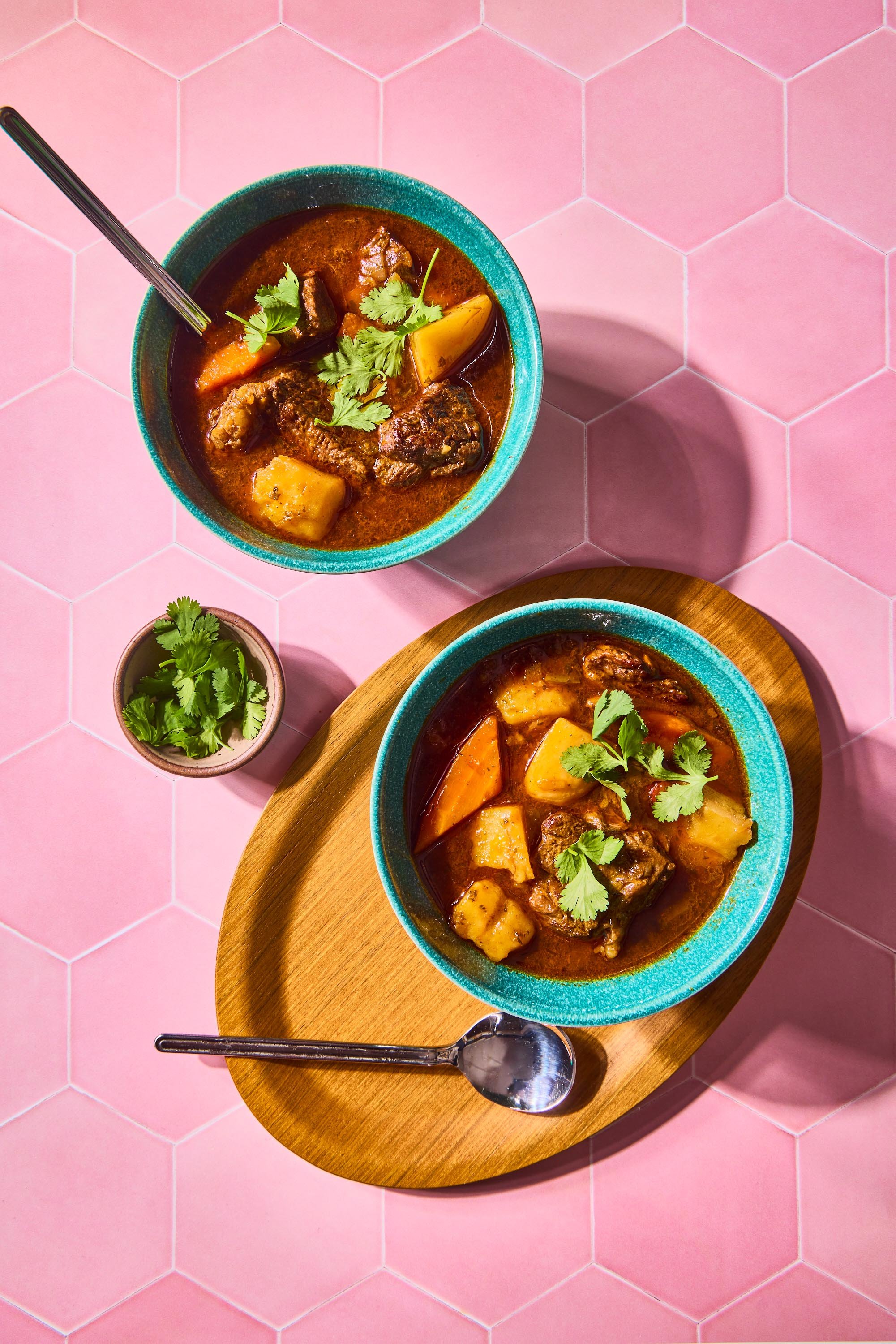 Two bowls of beef stew with potatoes and carrots, garnished with cilantro, on a pink tiled table with a small pot of cilantro and a wooden serving board.