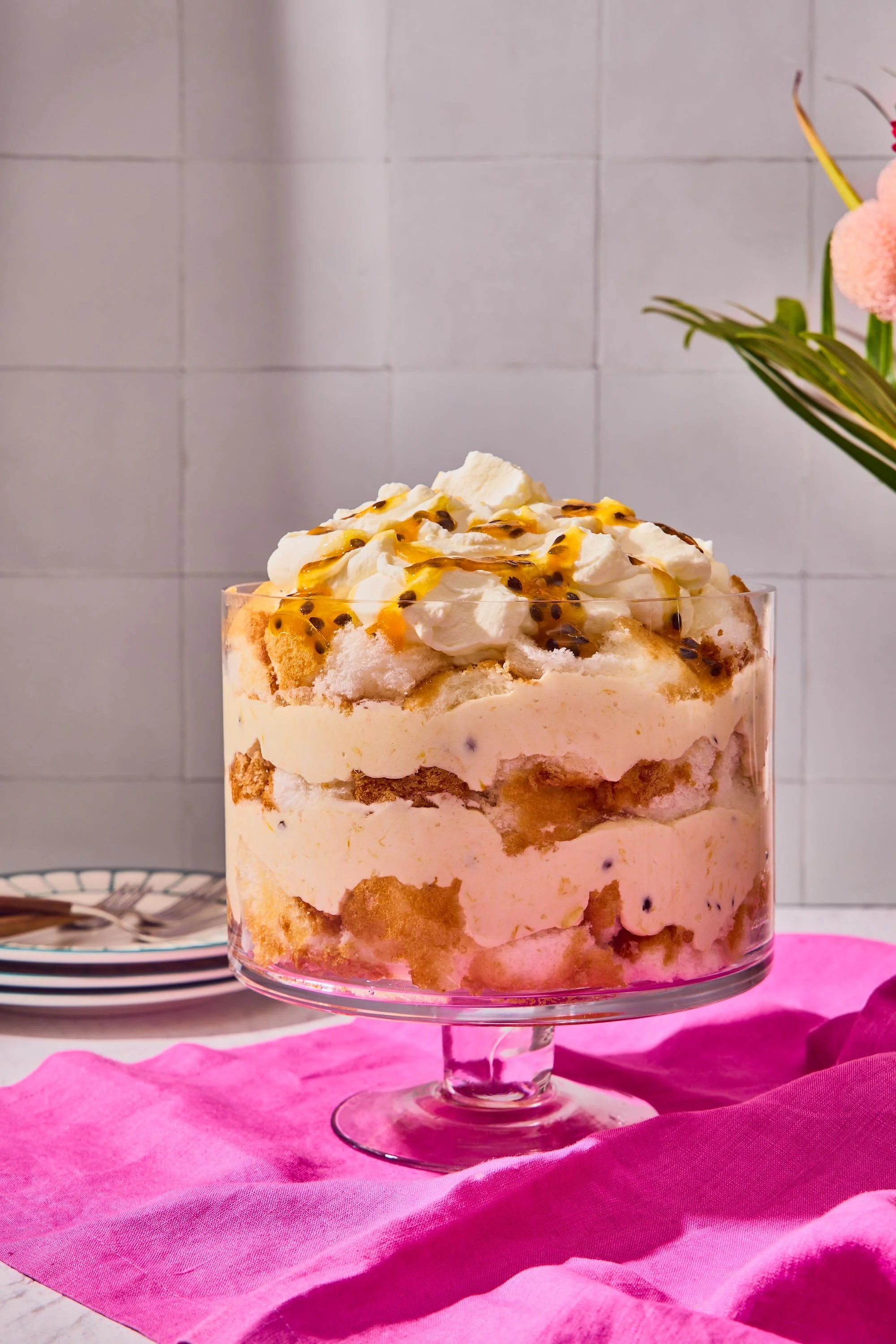 A layered dessert in a clear glass, topped with whipped cream and passion fruit seeds, placed on a pink cloth with a white tiled wall in the background.