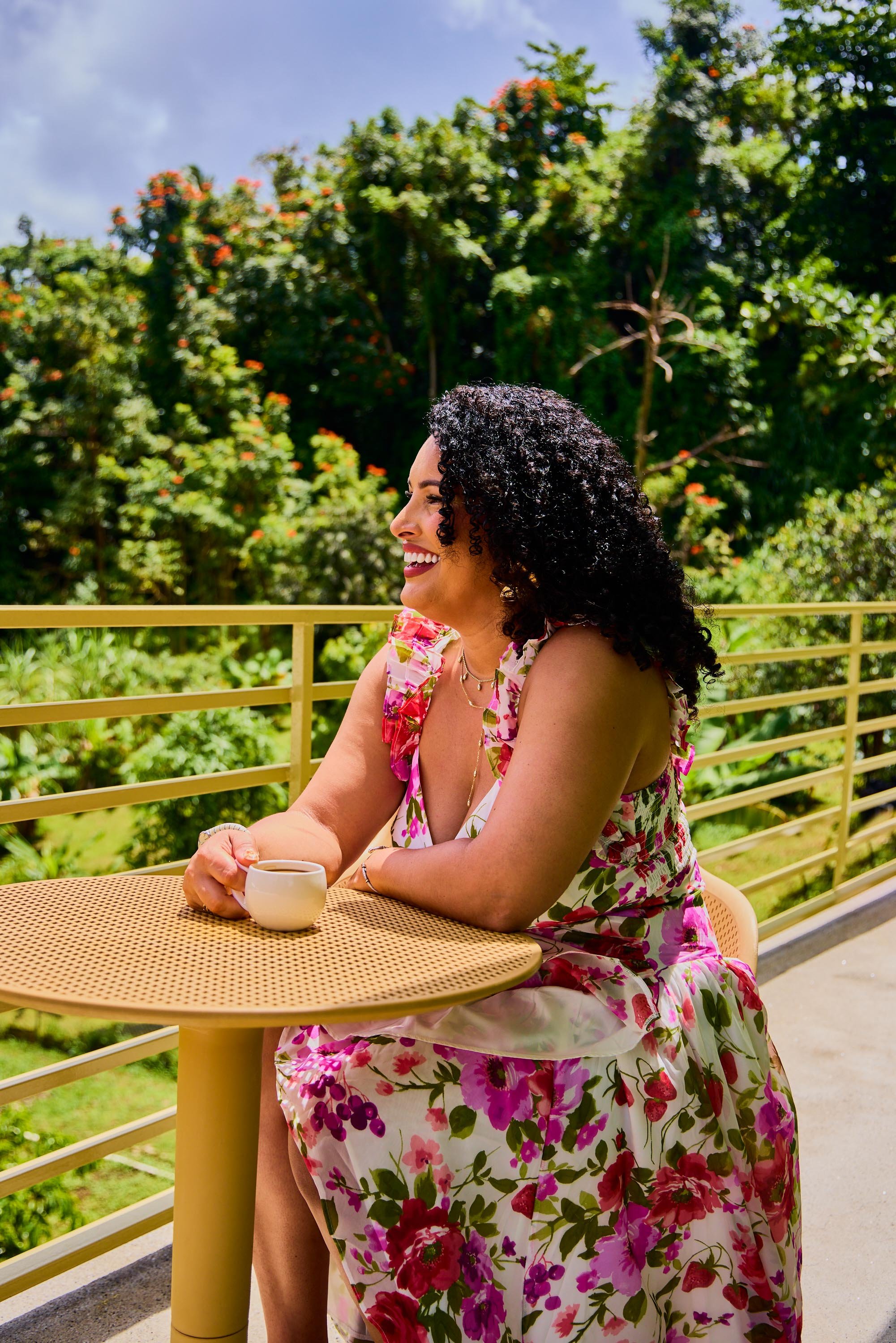 A woman with curly black hair smiling and sitting outdoors at a round yellow table, holding a white cup. She is wearing a colorful floral dress, and there are green trees in the background.