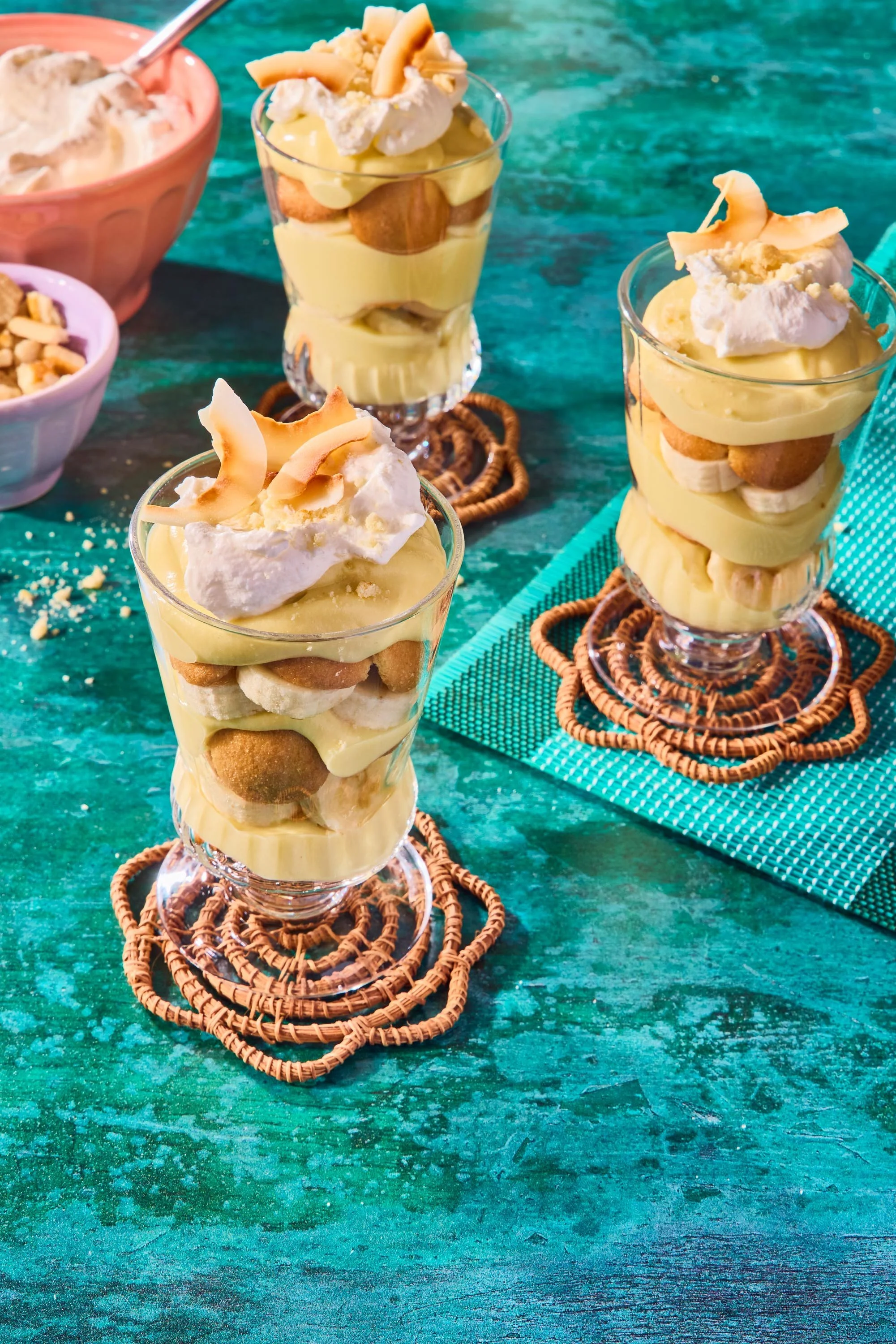Three glasses of banana pudding topped with whipped cream and coconut shavings, on a teal wooden table with woven coasters, with bowls of nuts and toppings in the background.