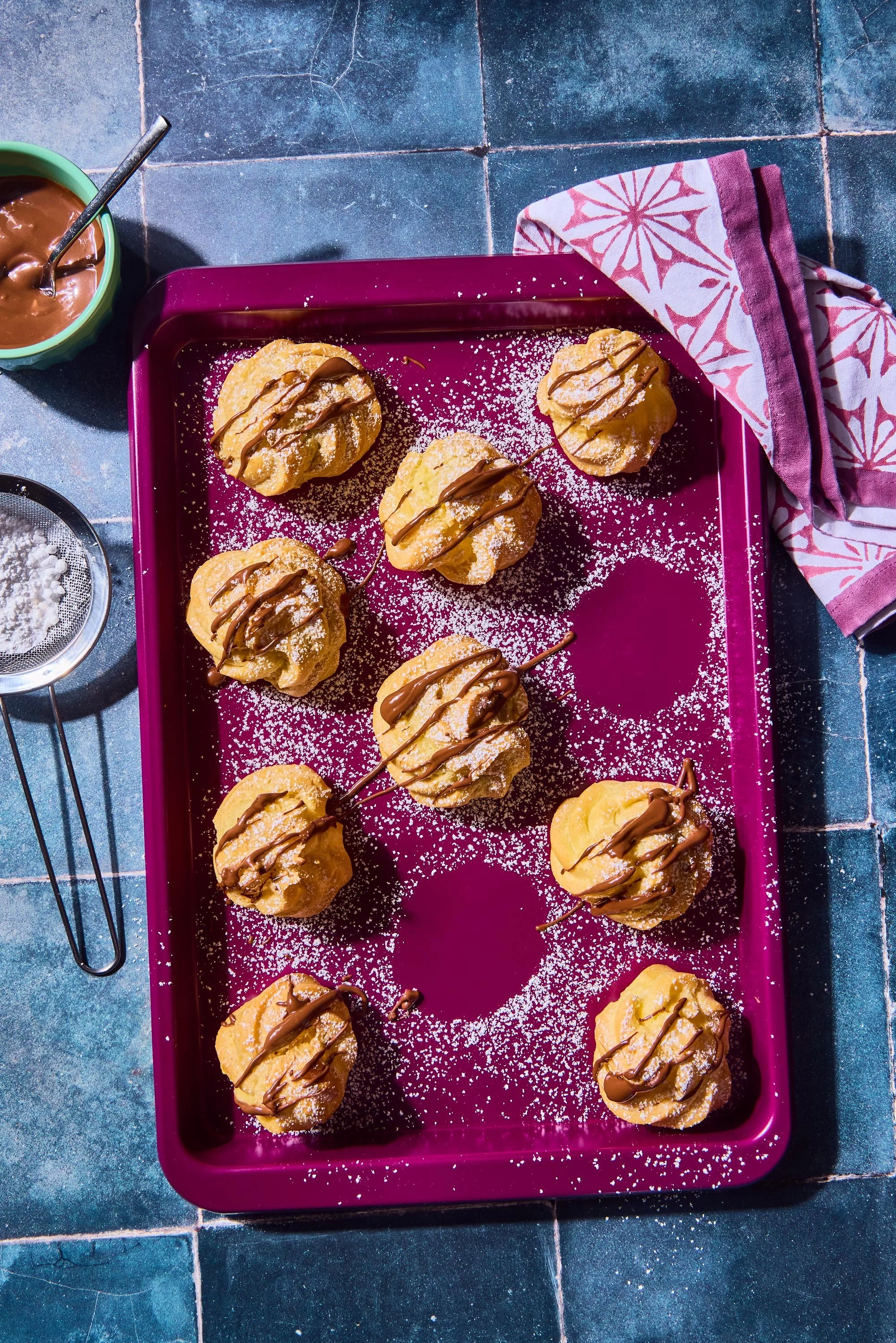 Cookies on a purple tray topped with powdered sugar and drizzled with chocolate, on a blue tiled surface with a pink and white cloth and a cup of chocolate