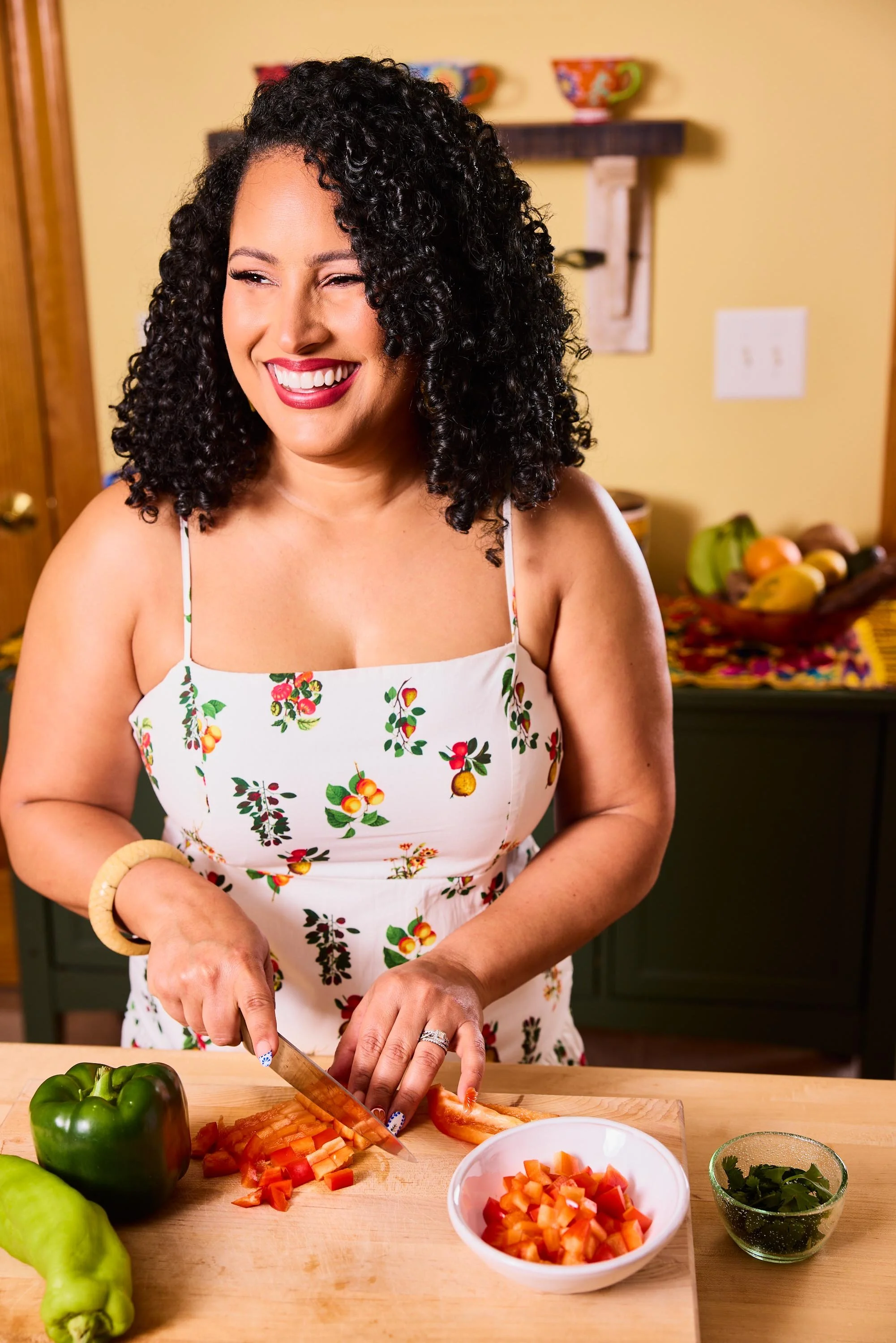 A woman with curly black hair and red lipstick chopping red bell peppers on a wooden cutting board in a kitchen. She is wearing a white dress with a cherry and lemon print, and a beige bracelet. There are green bell peppers on the counter, a bowl wit