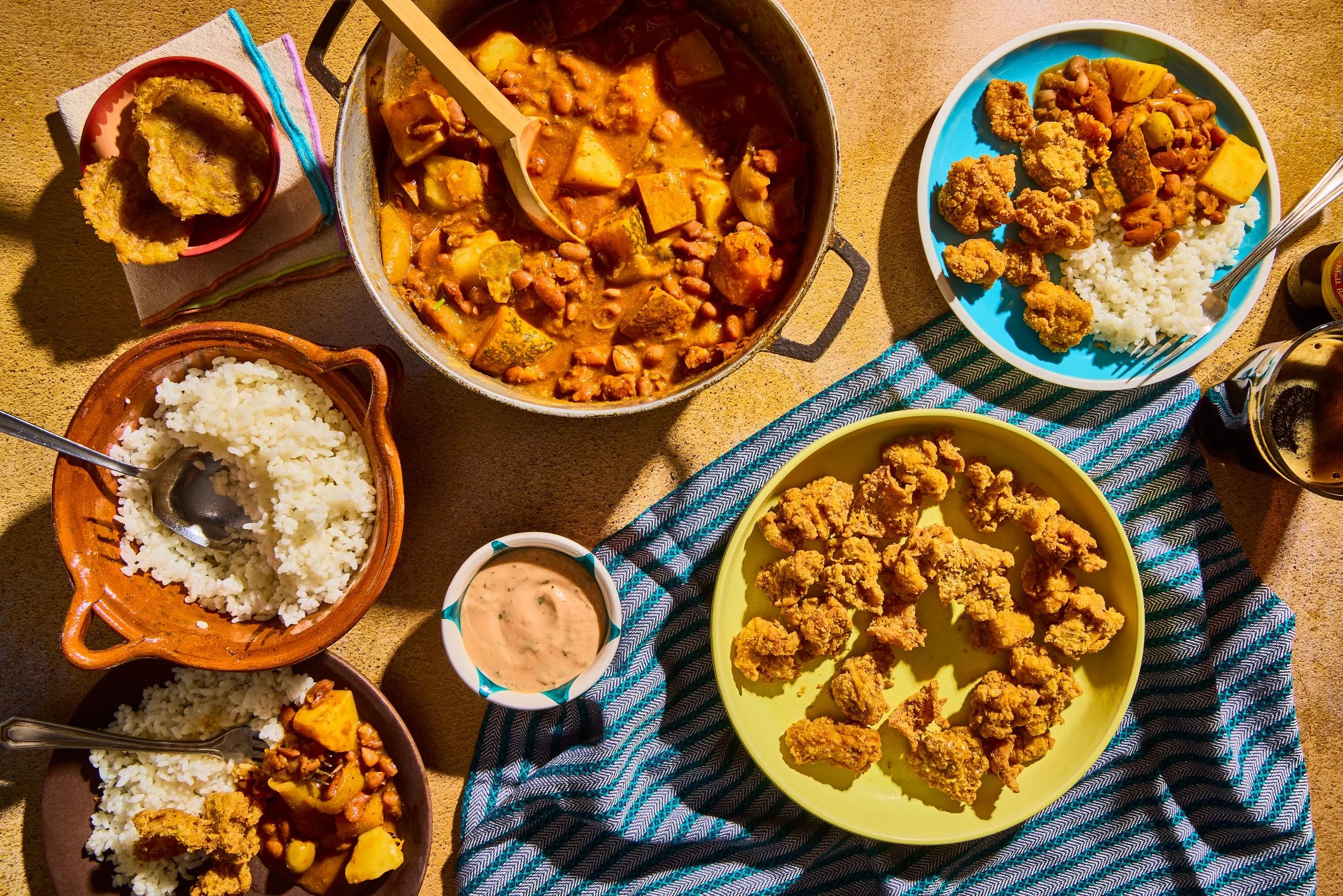 A top-down view of a table with various plates of food including fried chicken, white rice, a vegetable stew, and dipping sauces, set on a striped blue and white cloth on a sandy surface.