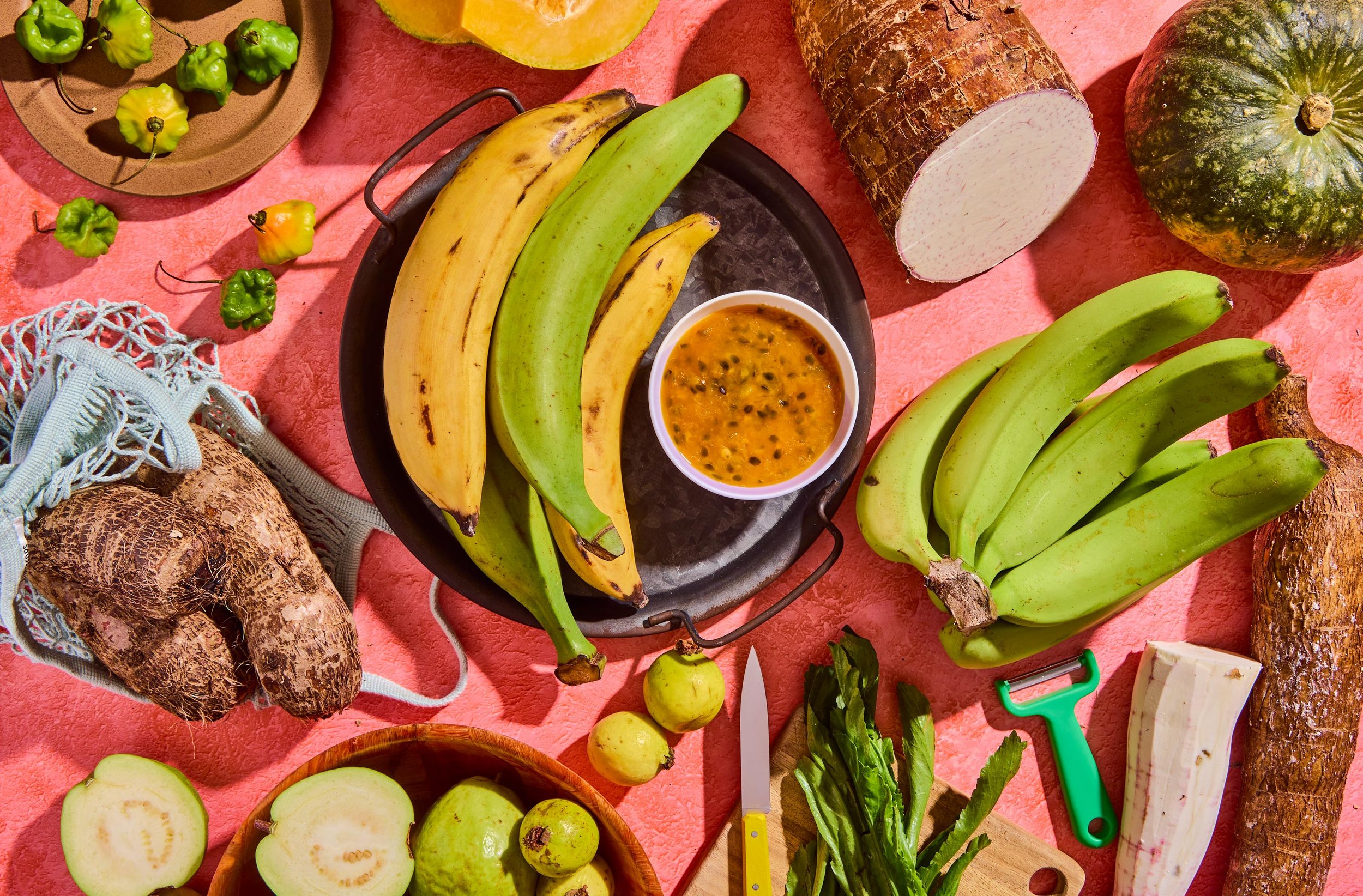 Assorted tropical fruits including bananas, jackfruit, green apples, and other produce on a pink surface with kitchen tools and a bowl of passion fruit pulp.