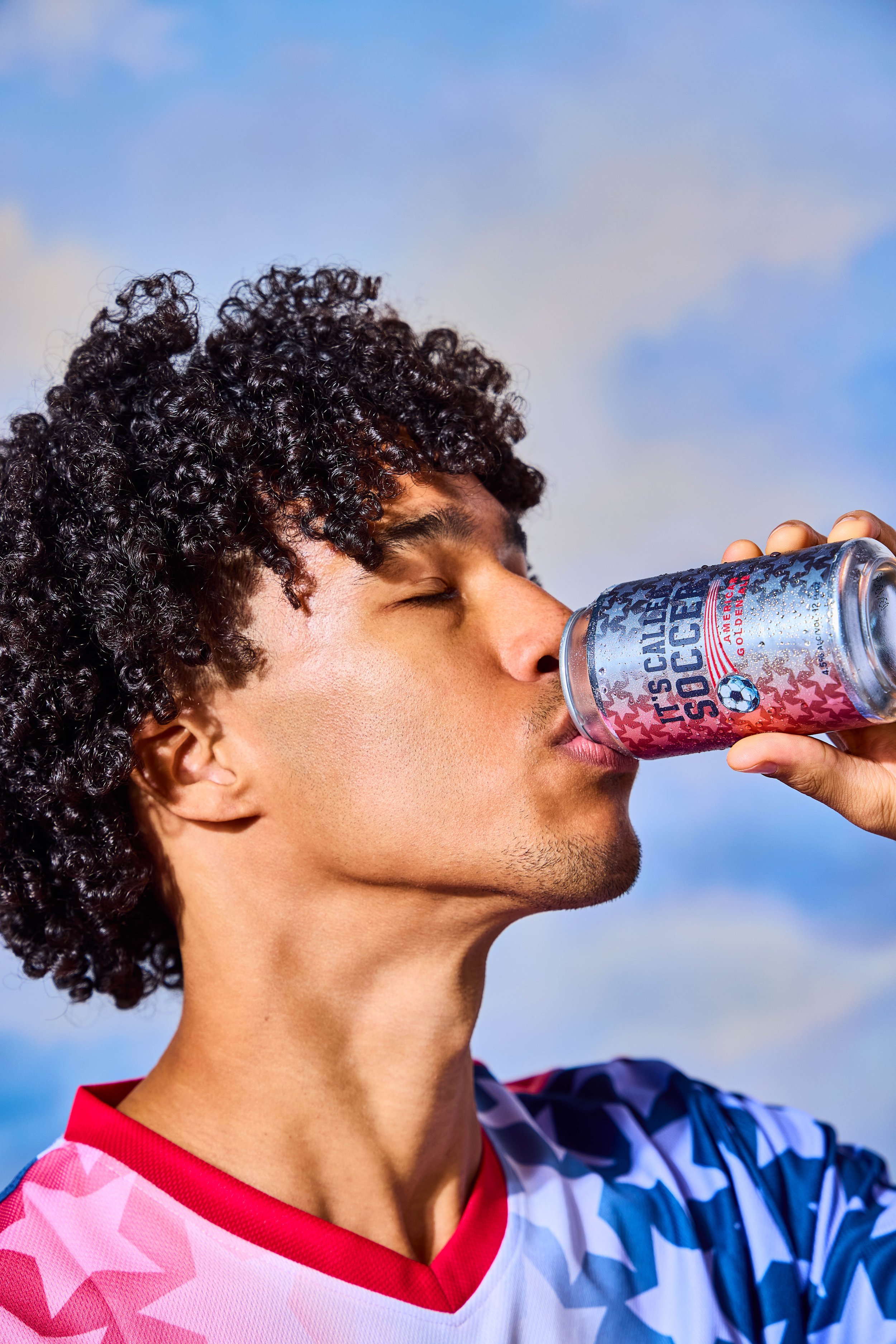 A young man with curly hair wearing a sports jersey, drinking from a clear cup with a patriotic design that says 'It's Caller Go' and a soccer ball, outdoors with blue sky and clouds.