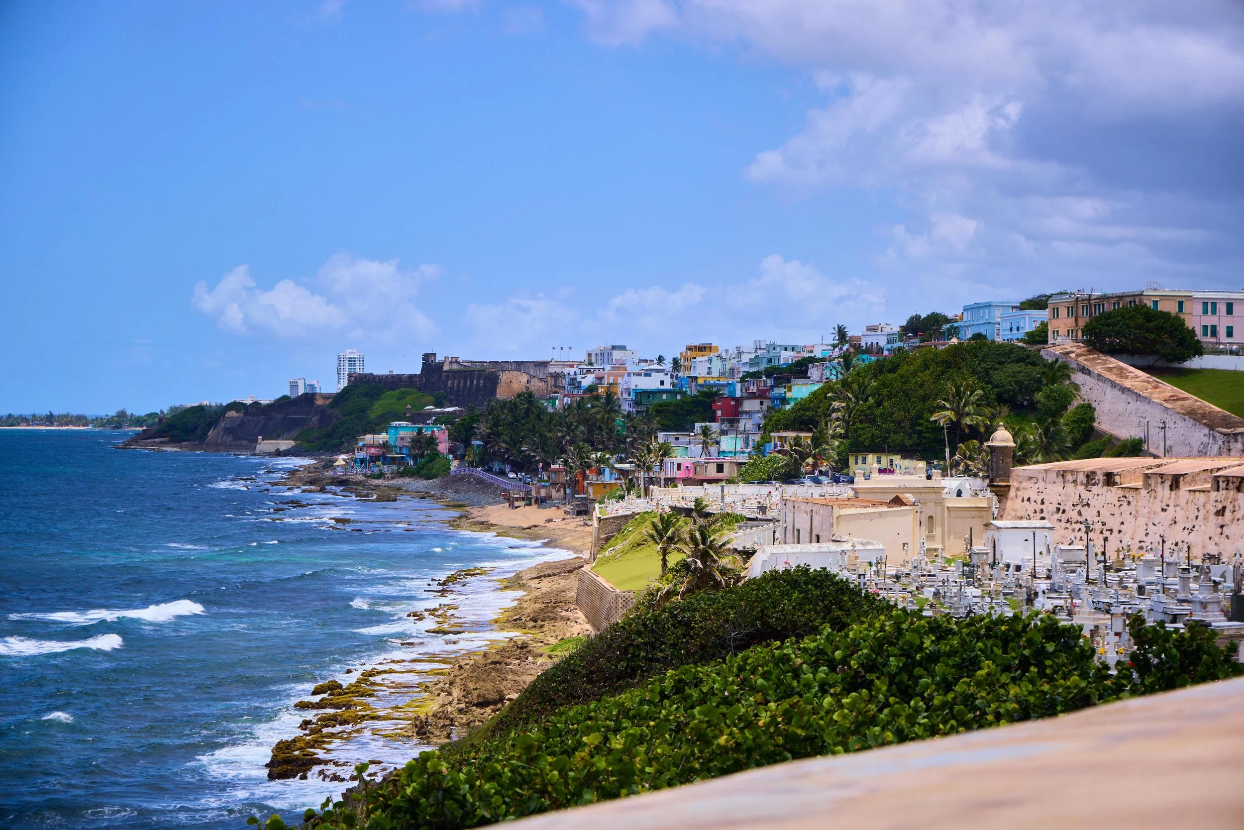 A coastal town with colorful buildings on a hillside overlooking the ocean. There is a cemetery with white tombstones in the foreground and a blue sky with some clouds.