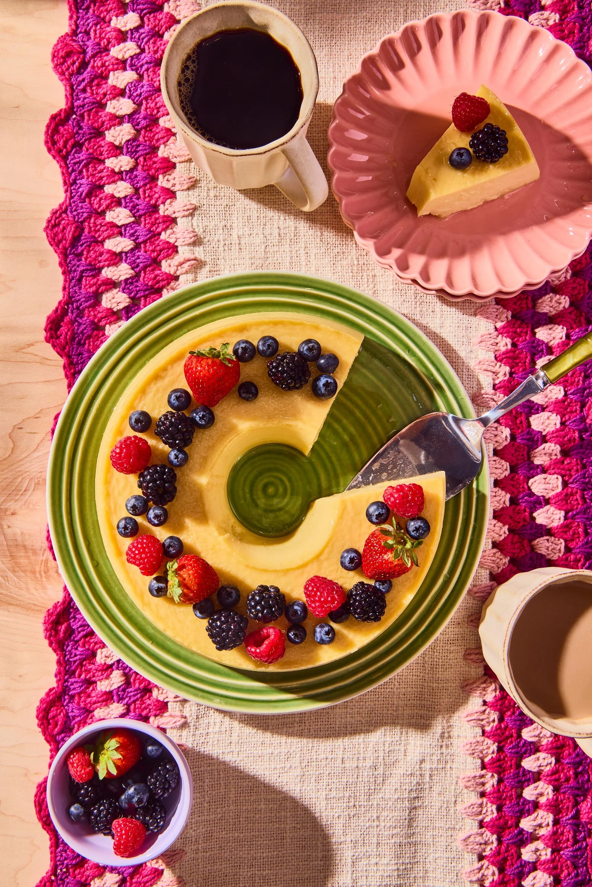 A table with a yellow cheesecake topped with mixed berries on a green-rimmed plate, a pink dish with a slice of cheesecake topped with berries, a cup of coffee, and a small bowl of mixed berries, all on a woven pink and beige table runner.
