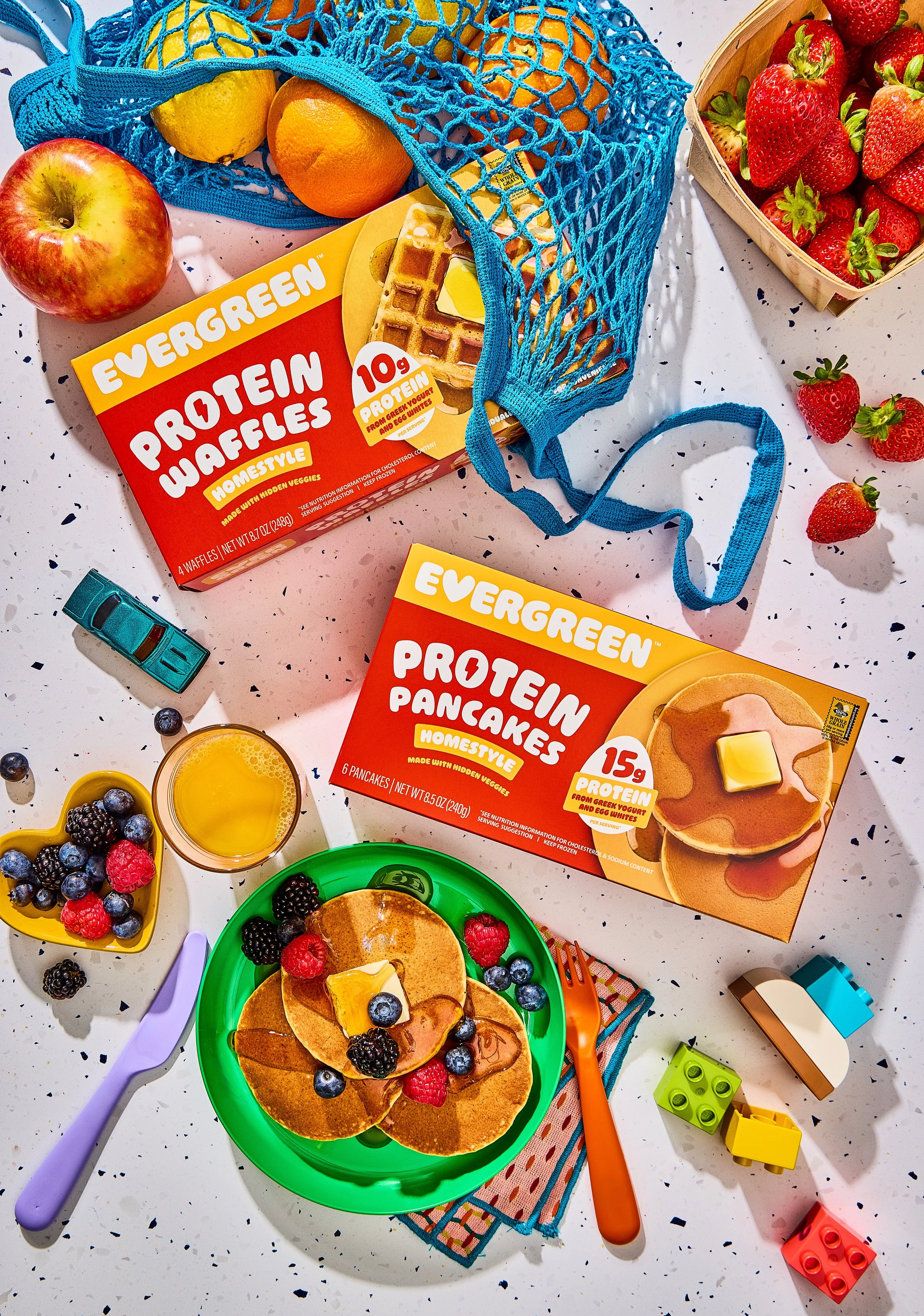A breakfast scene with pancakes topped with blueberries and raspberries, a glass of orange juice, and bowls of mixed berries on a speckled white table. There are kitchen toys and utensils around, along with superhero-themed pancake boxes.