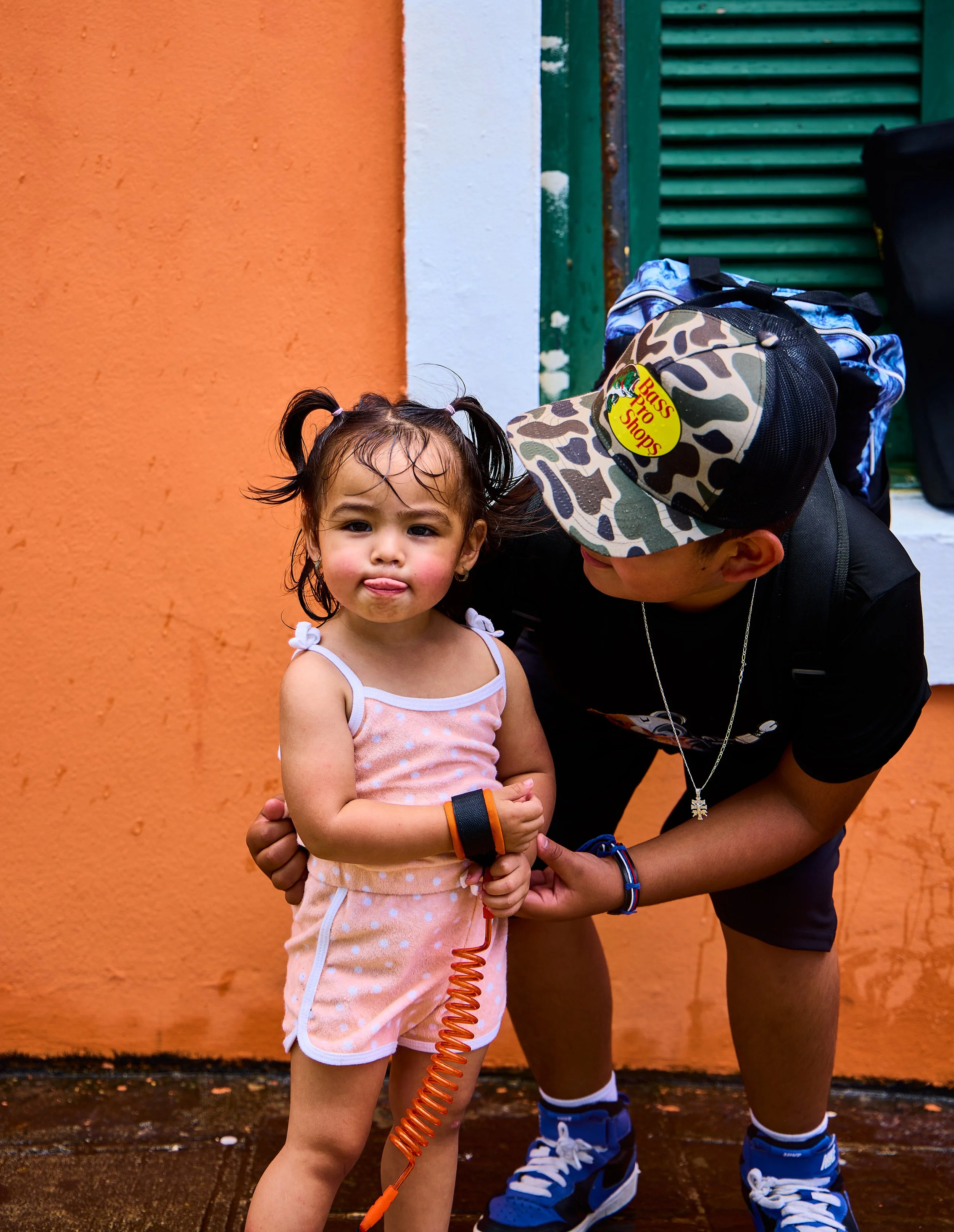 A young girl with pigtails and a pink polka dot dress standing outdoors against an orange wall, with a man wearing a camouflage cap, black shirt, and blue sneakers.