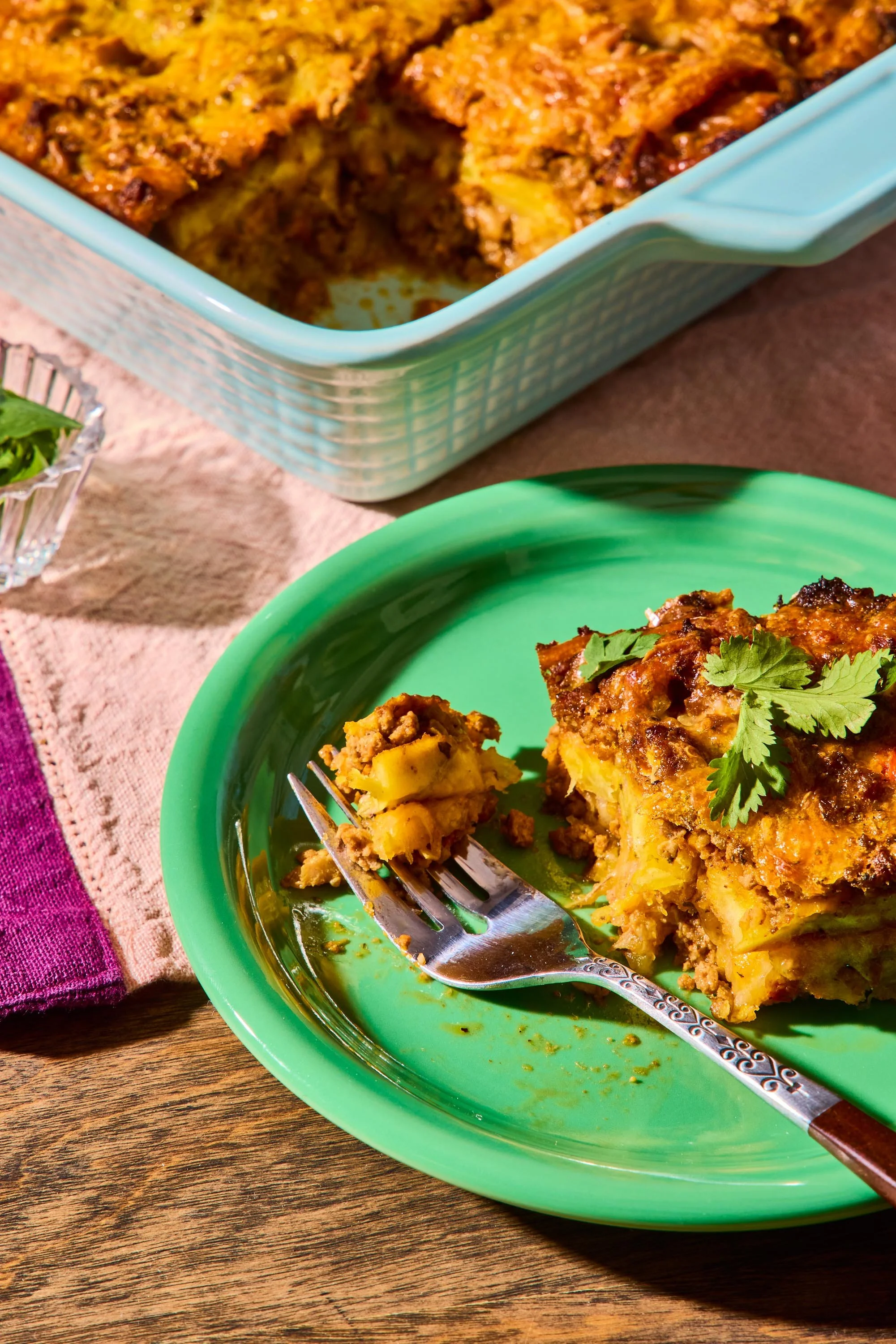 A serving of baked casserole with ground meat, potatoes, and seasonings on a green plate garnished with cilantro, with a piece on a fork. In the background, a blue dish contains more casserole, on a wooden surface with a pink and purple cloth.