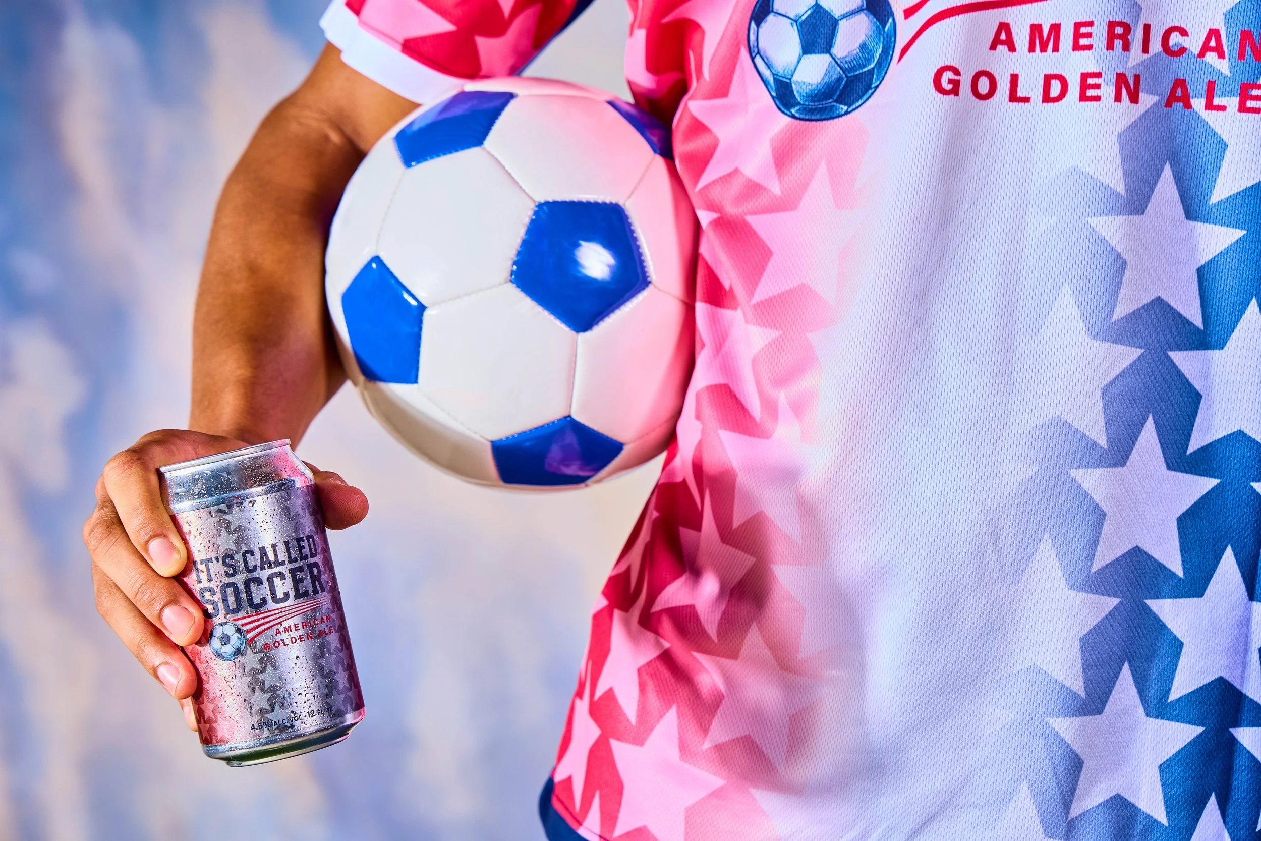 Person wearing a festive patriotic soccer jersey holding a blue and white soccer ball in one arm and a silver can of soda in the other.