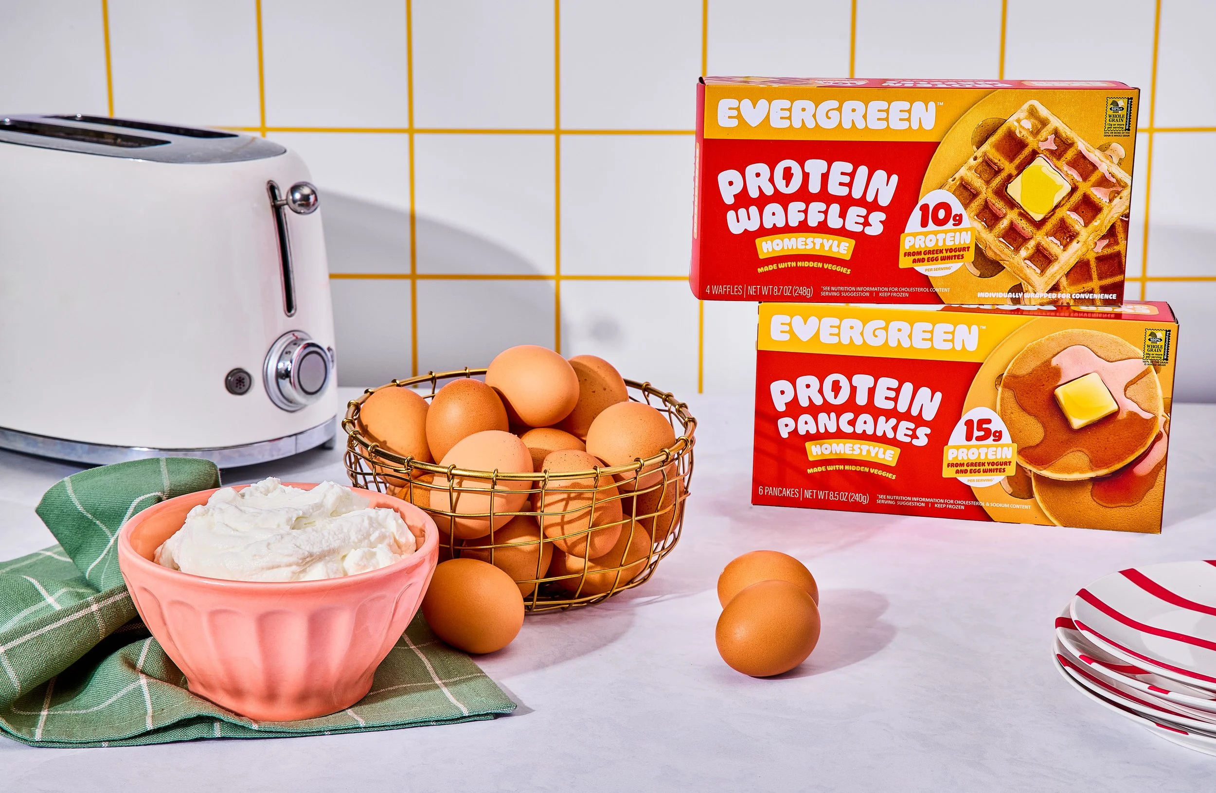 A kitchen counter with a white toaster, a basket of eggs, a bowl of whipped cream, several plates, and two boxes of protein waffles and pancakes on a tiled yellow and white wall background.