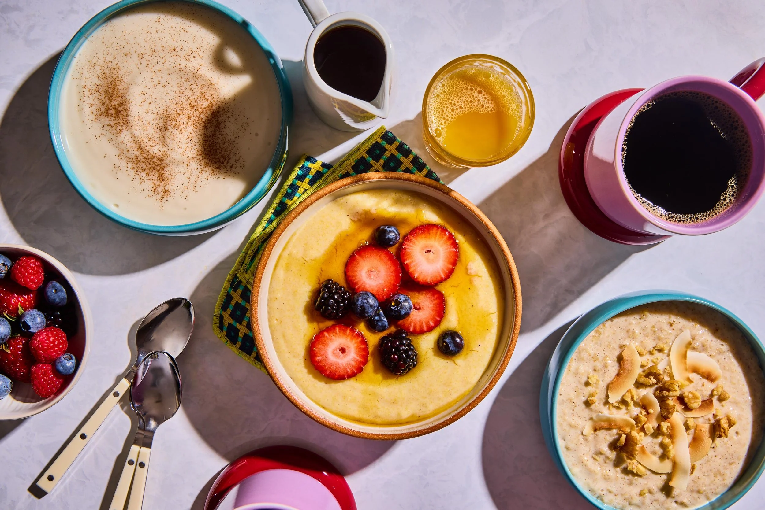 Assorted bowls of dessert, including pudding topped with strawberries and blueberries, a creamy custard, and a chocolate cake, with coffee and syrup on a white surface.