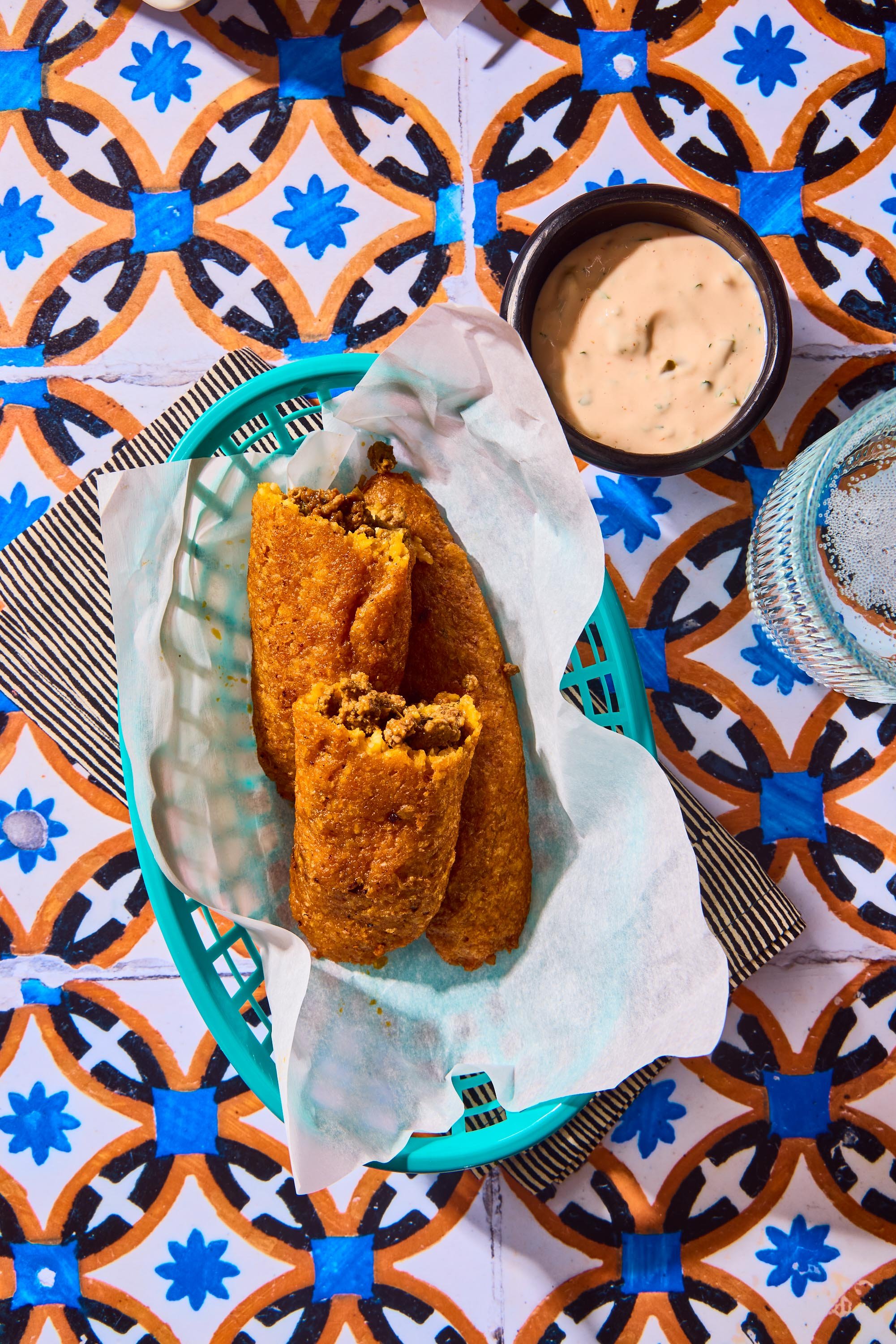 Fried taquitos served on a paper-lined basket, accompanied by a cup of creamy dipping sauce, on a colorful tile table.