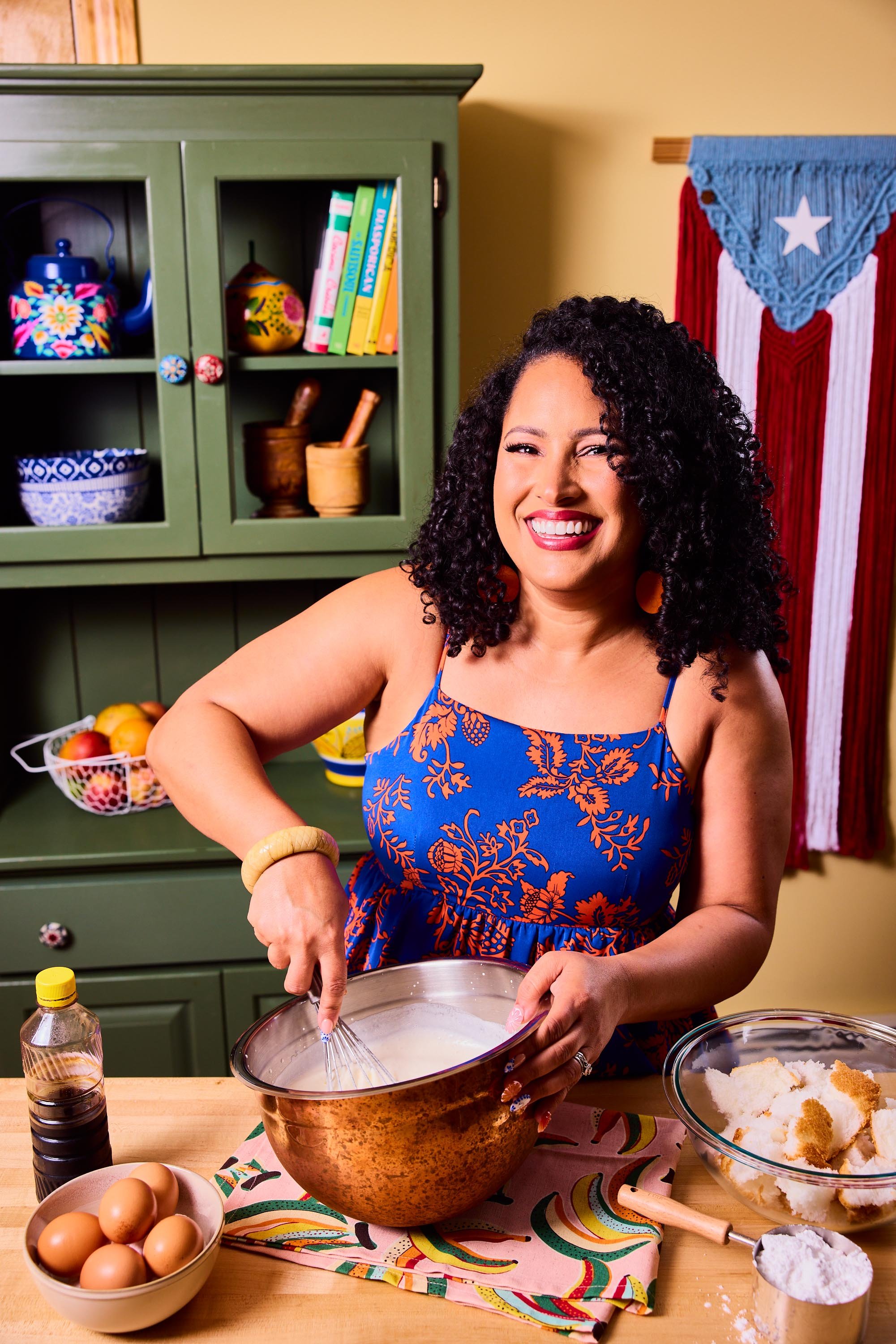 A woman with curly black hair wearing a blue and orange floral dress mixing ingredients in a metal bowl in a colorful kitchen.