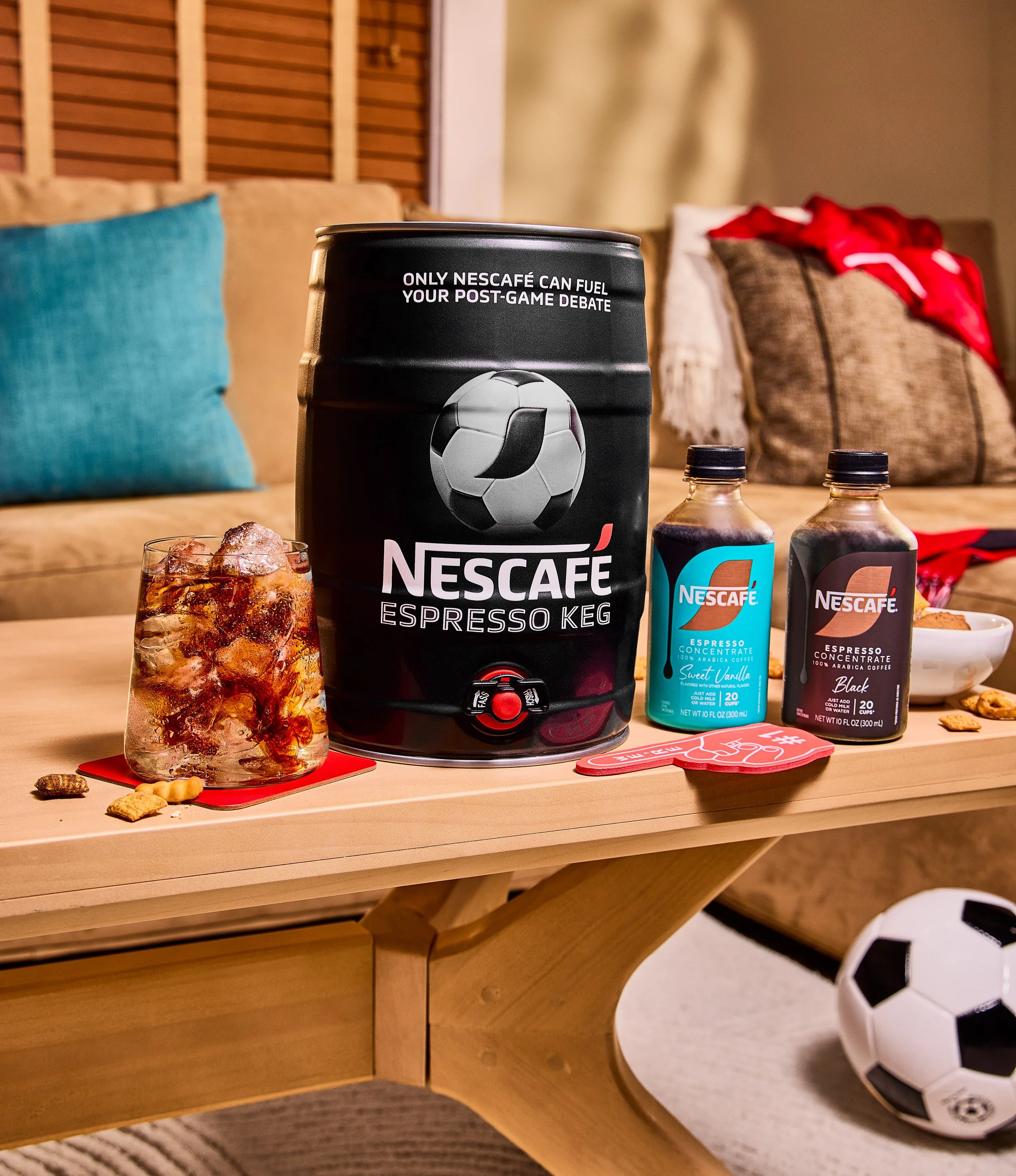 A can of Nescafé espresso coffee, a glass of dark iced coffee with ice cubes, two bottles of flavored syrup or concentrate, a small bowl of snacks, a red coaster, and a soccer ball on a wooden table, with a couch and pillows in the background.