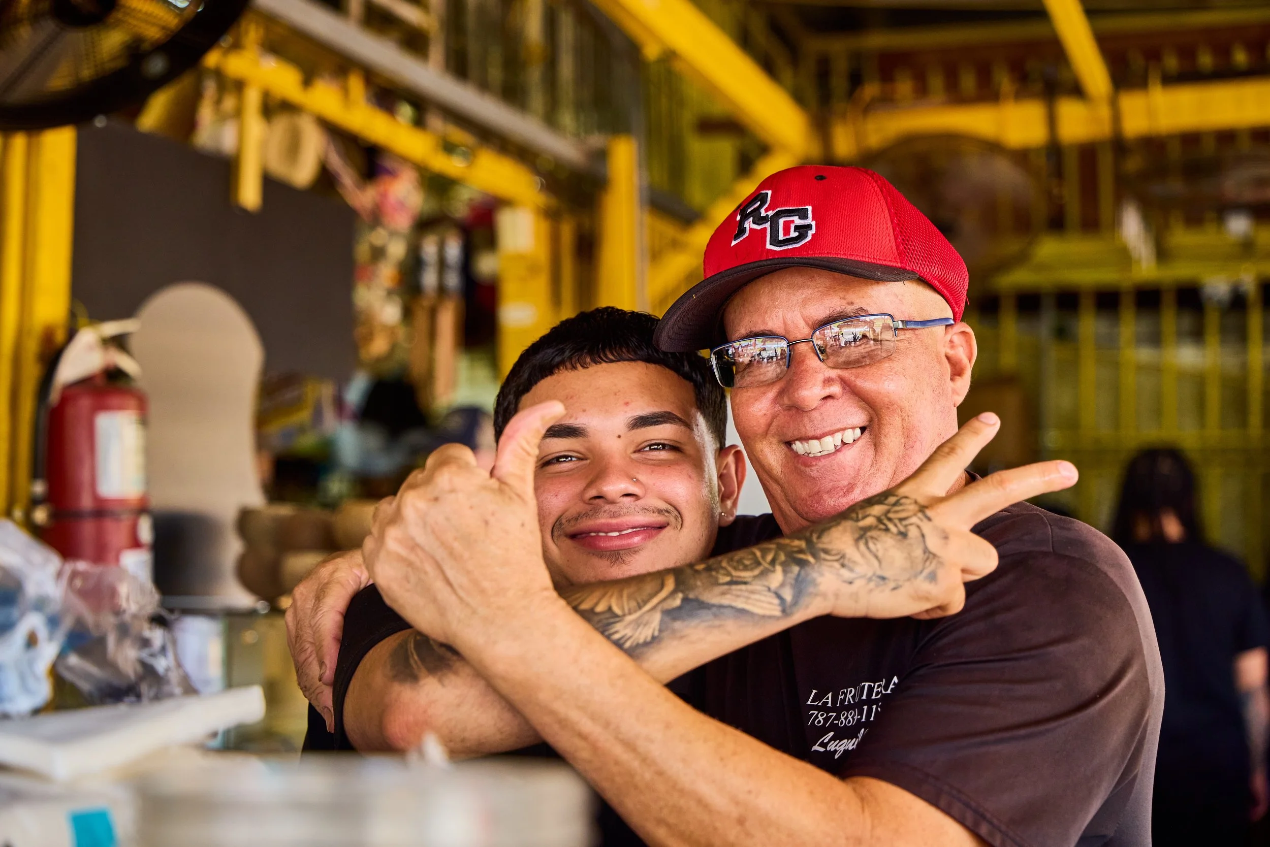 Two men hugging and smiling in a workshop or hardware store with shelves and a fire extinguisher in the background.