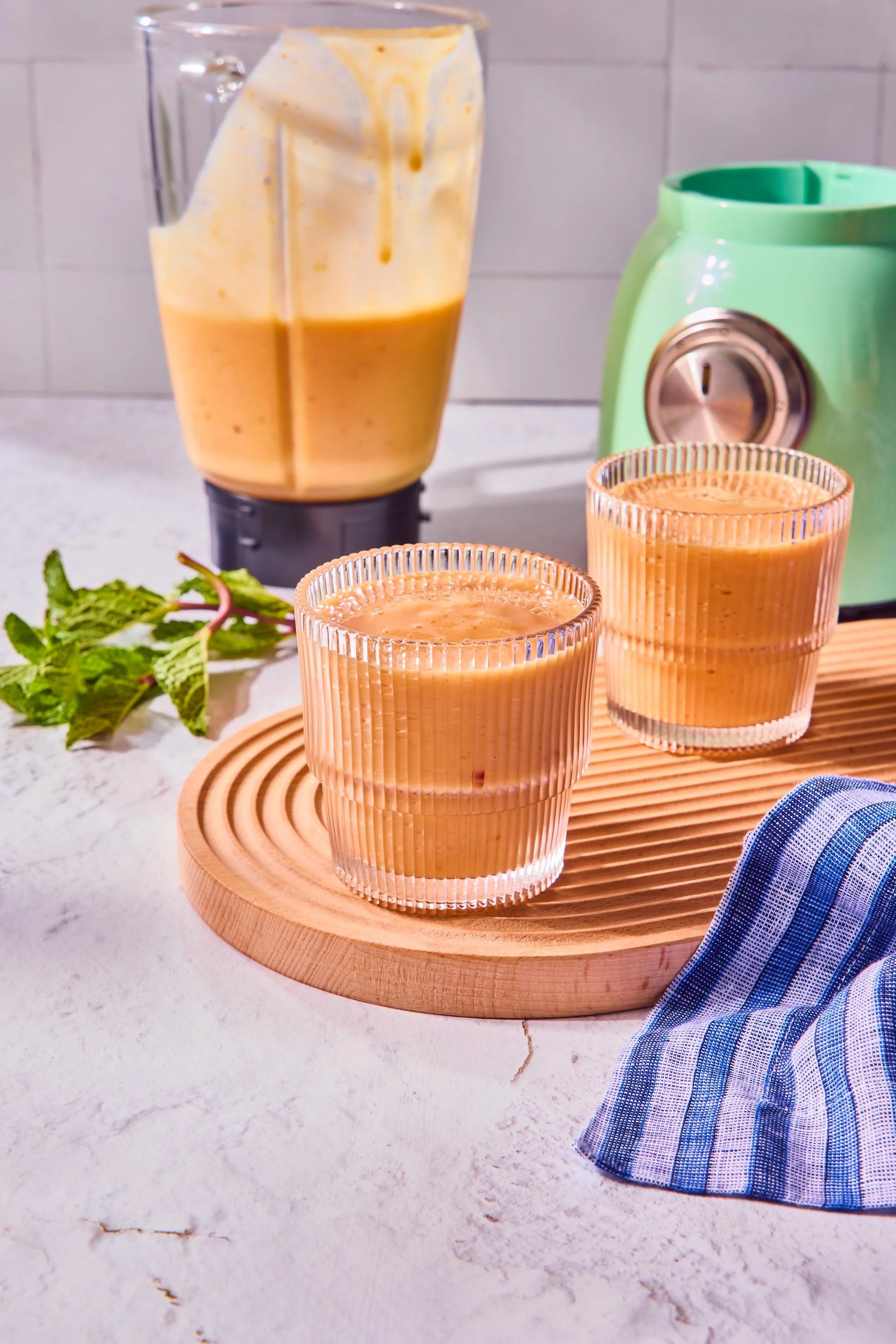 A blender with a creamy orange-colored smoothie, two glasses filled with the same smoothie on a wooden tray, fresh mint leaves on the white countertop, and a blue striped cloth nearby.