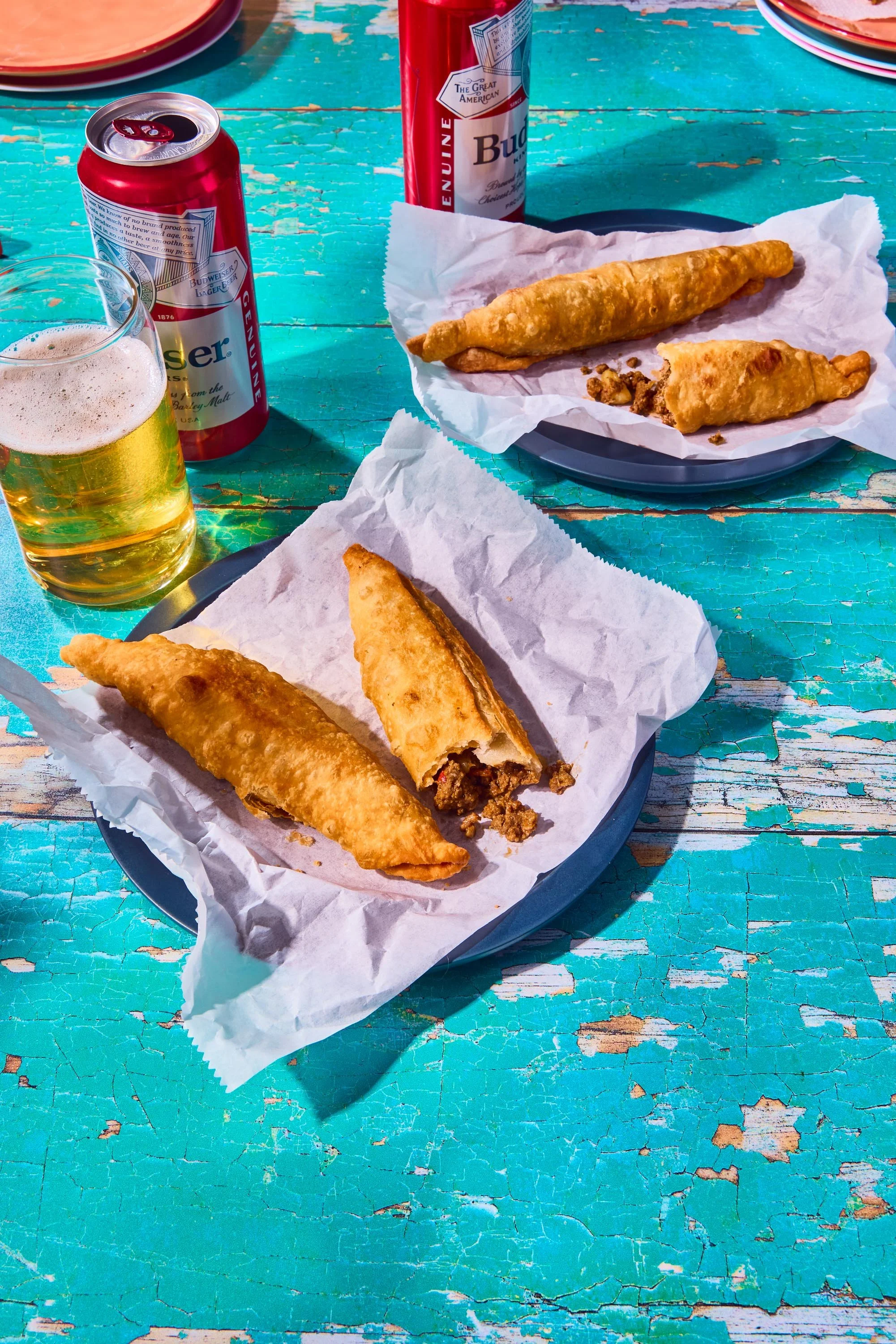 Fried fish fillets on plates, two cans of Budweiser beer, and a glass of beer on a weathered turquoise table.