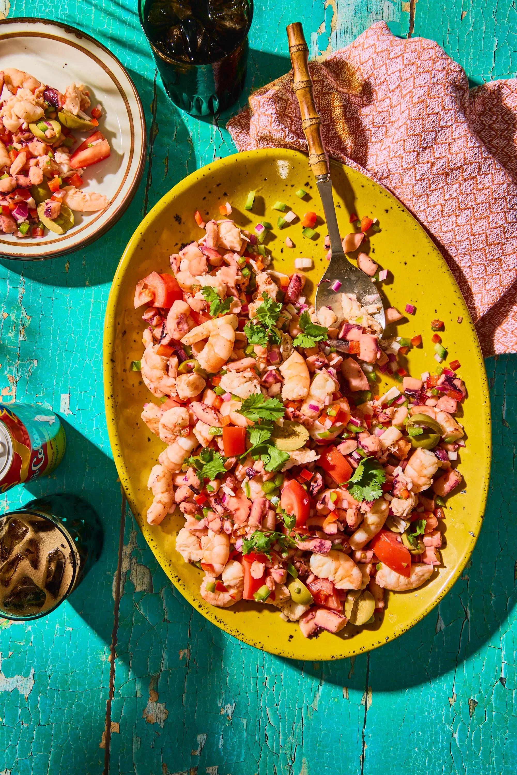 A colorful outdoor meal setup featuring a large yellow plate with a seafood salad garnished with cilantro, a smaller plate with more seafood salad, and three glasses of soda on a weathered turquoise wooden table, with an orange and white patterned cl