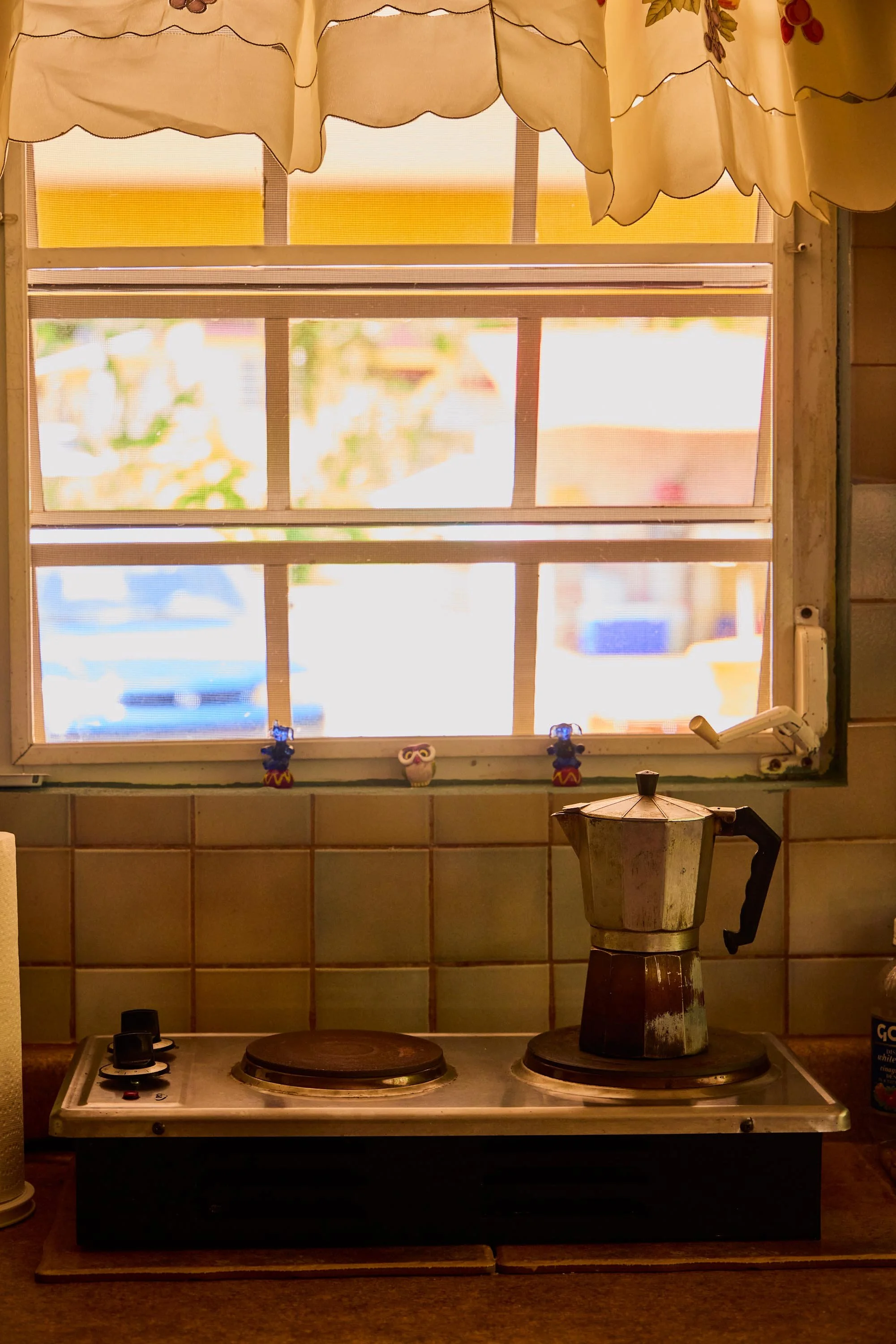 A kitchen window with a yellow valance, a stovetop with an electric burner, and a moka pot on the stove. The window has a screen and small decorative figurines hanging from the window frame.
