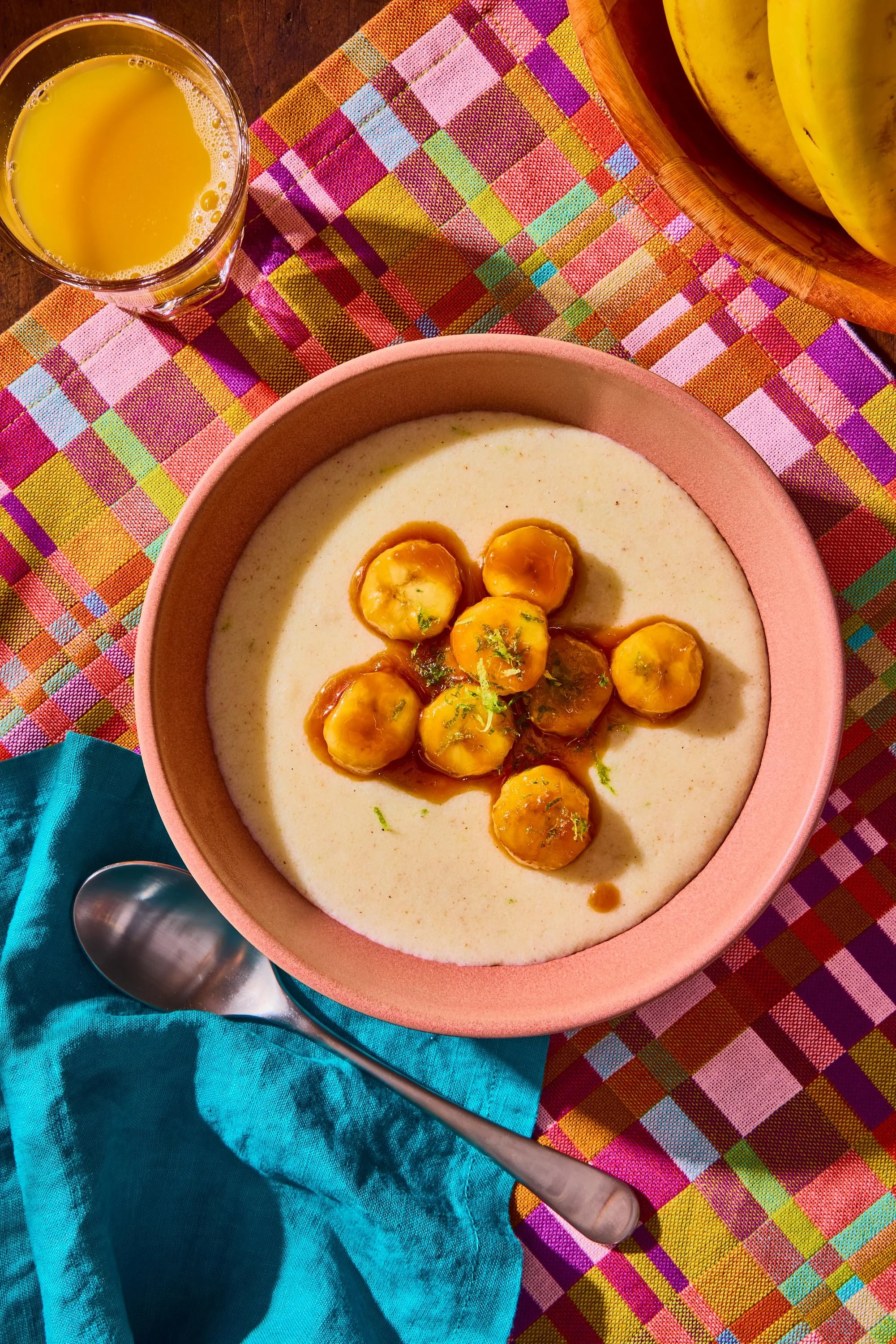A bowl of creamy soup topped with small yellow dumplings, a glass of orange juice, and a banana on a wooden table with colorful tablecloth and a turquoise cloth.