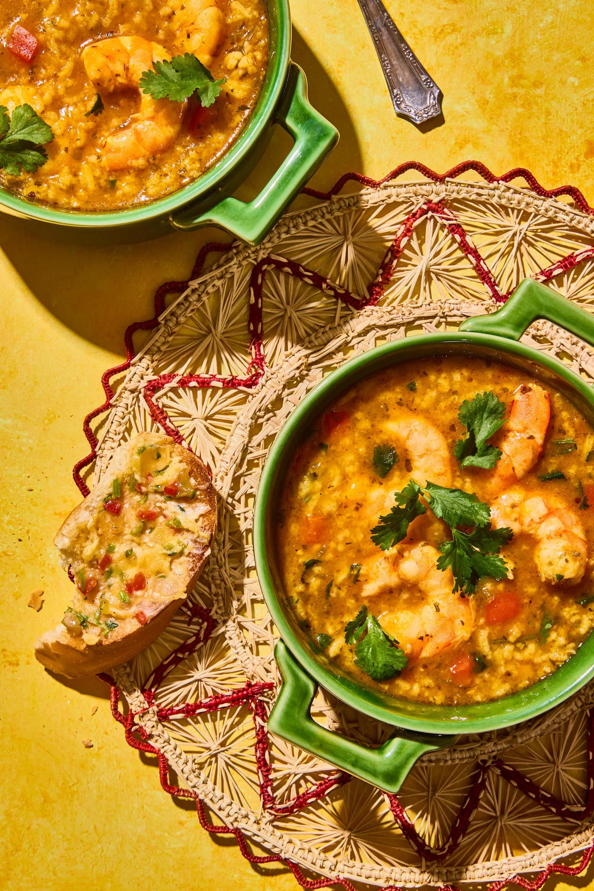 A bowl of shrimp and vegetable soup garnished with cilantro on a yellow surface, along with a piece of garlic bread topped with cheese and red and green chopped peppers.