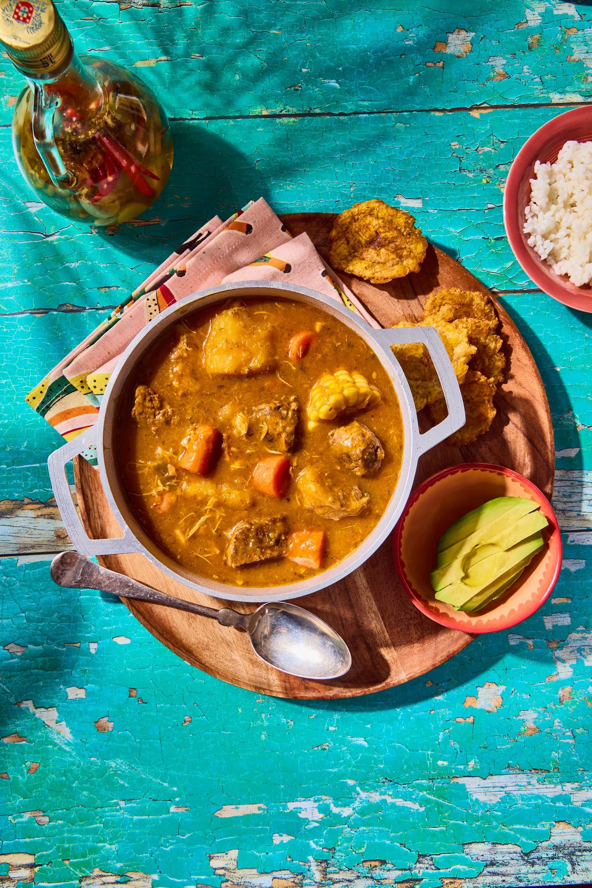 A bowl of hearty stew with vegetables and chunks of meat, accompanied by fried plantains, a small bowl of sliced avocado, white rice in a pink bowl, and a glass bottle of oil on a distressed turquoise wooden table.