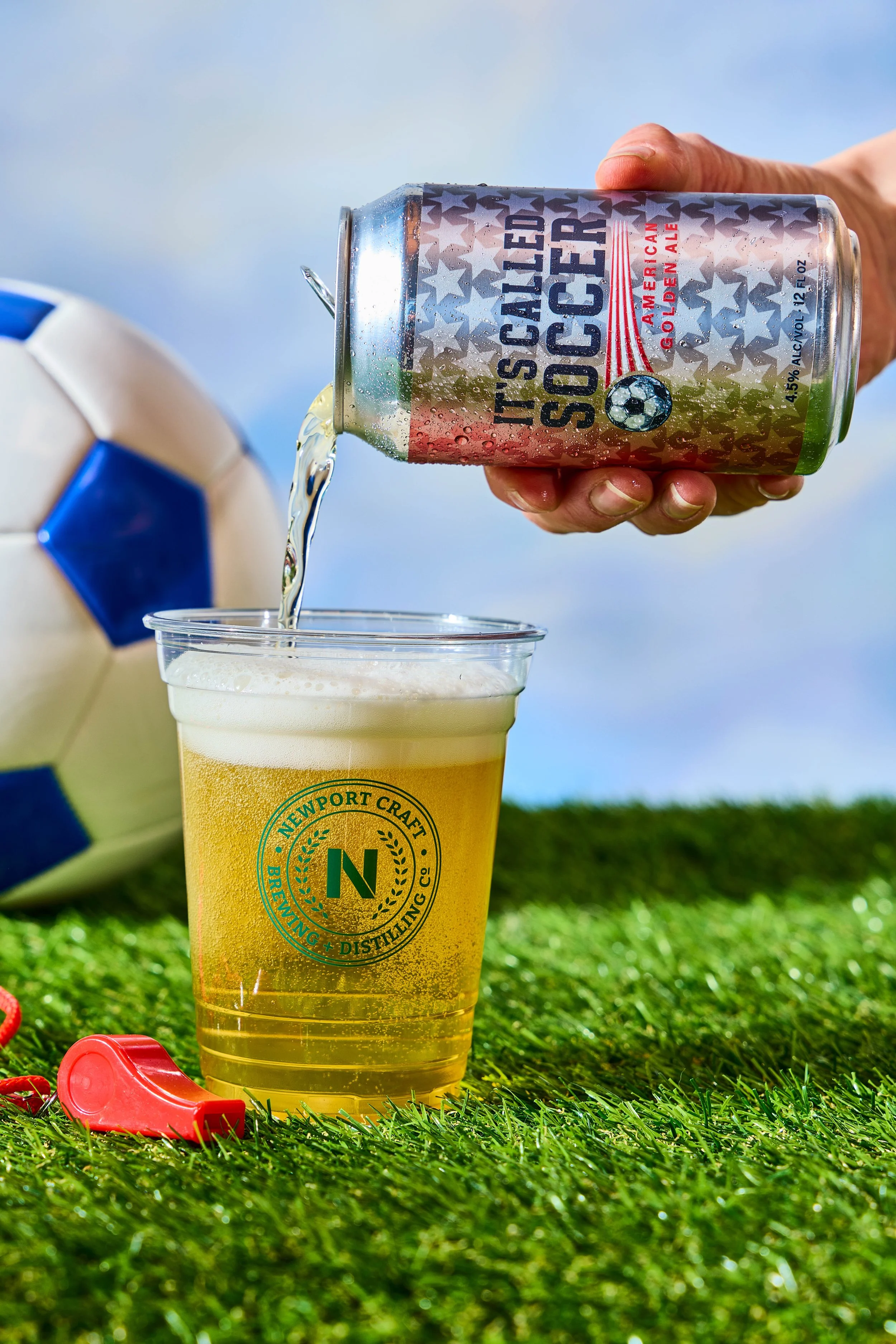 A person pours a can of American Golden Ale into a plastic cup with brewery branding, on a grass field next to a soccer ball and a red whistle, under a blue sky.