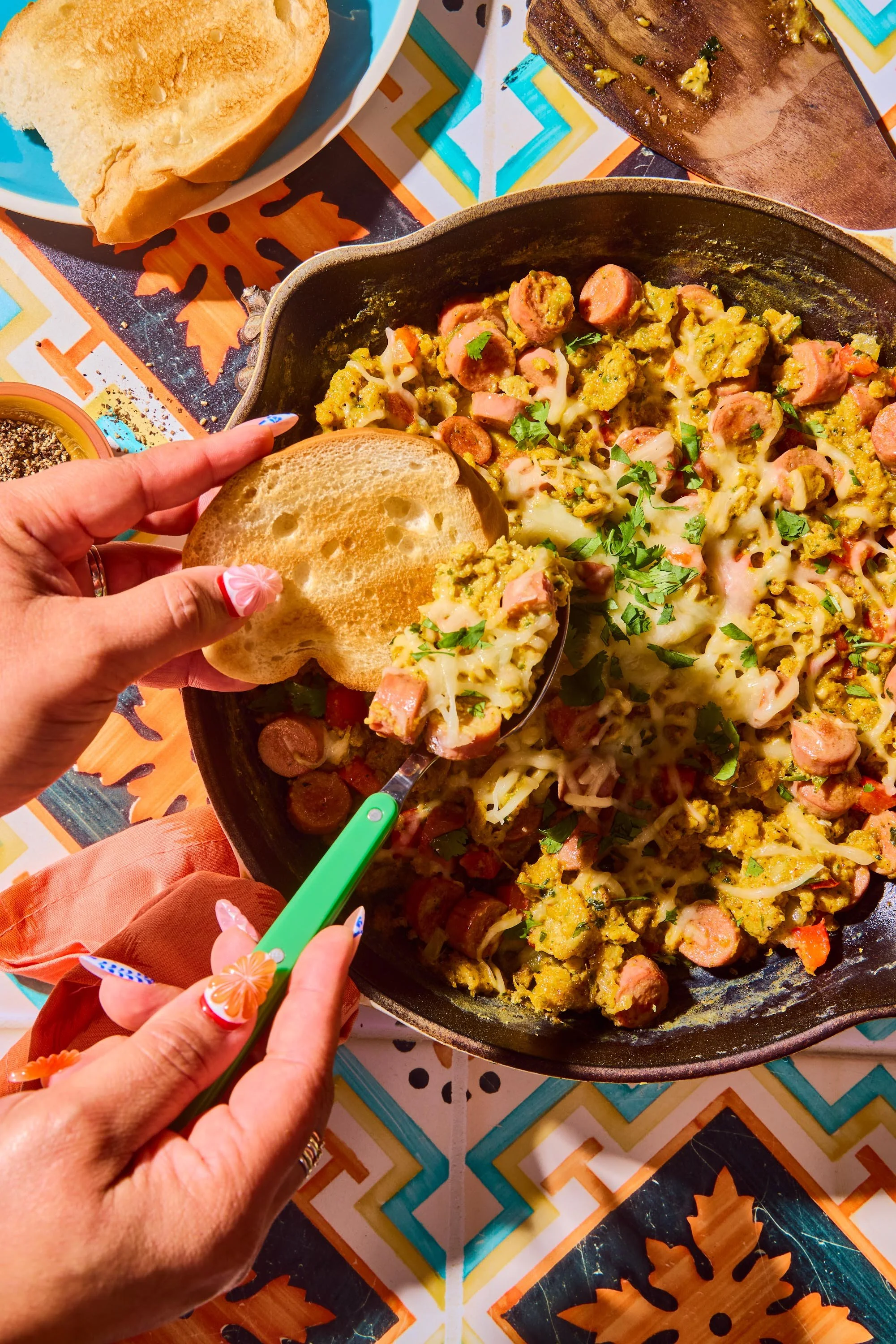 A close-up of a skillet filled with a mixture of cooked sausage, scrambled eggs, and cheese, garnished with chopped herbs. A hand is holding a piece of toasted bread and a green utensil is also seen. The background shows a table with colorful geometr