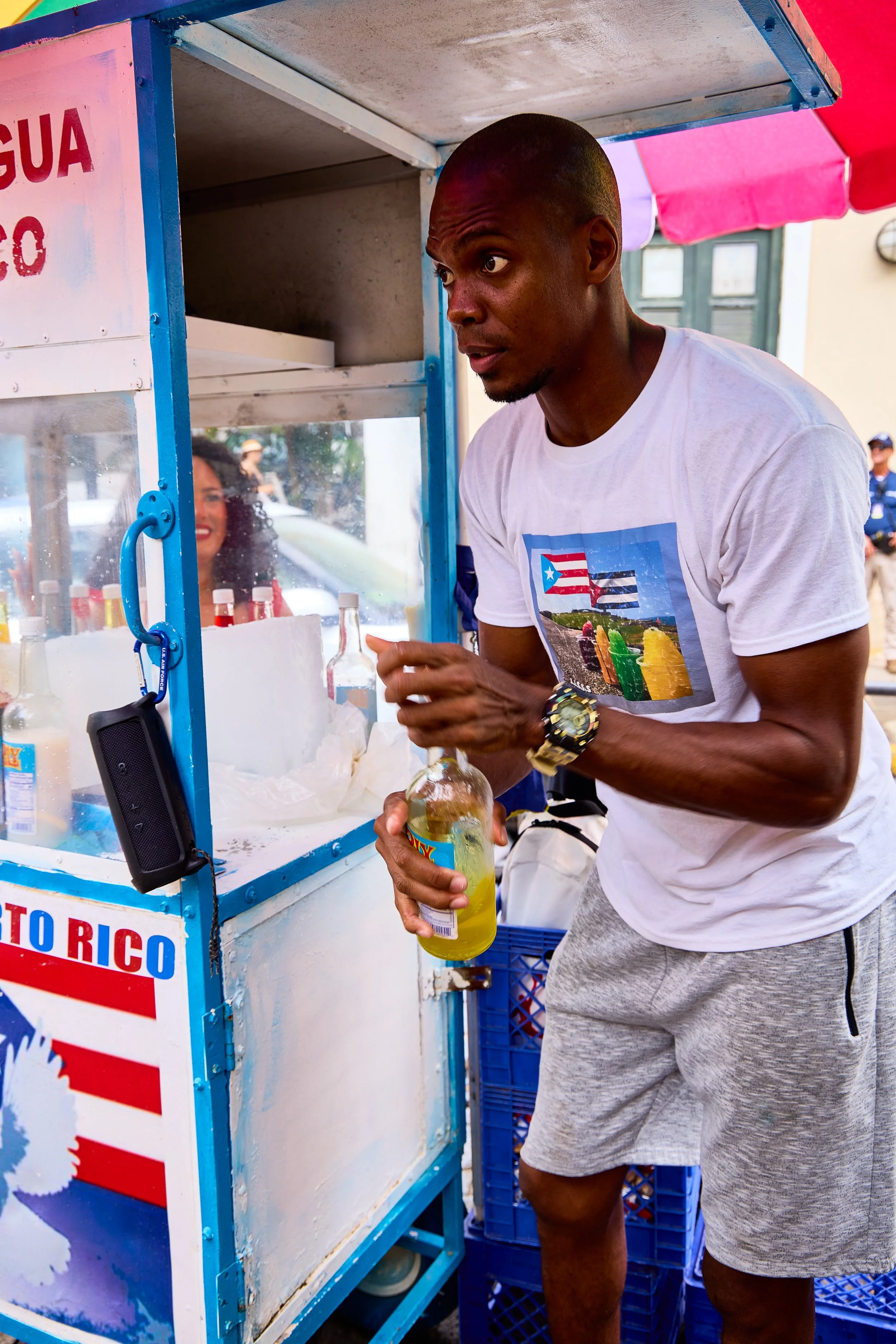 A man in a white t-shirt and gray shorts buying a yellow beverage from a street vendor's cart with bottles of syrup and a woman smiling inside the cart.