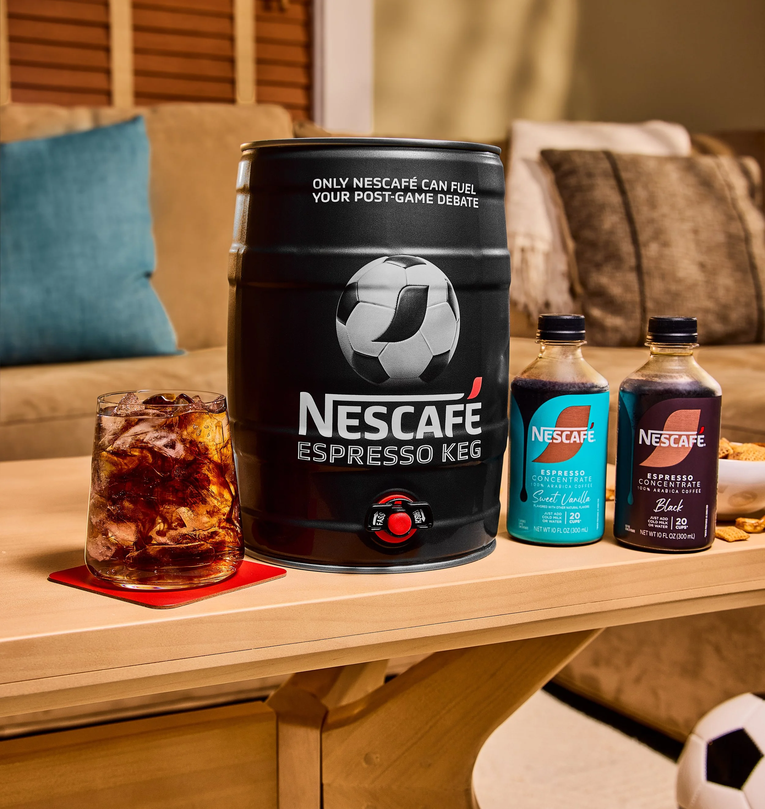 A black NESCAFÉ espresso coffee keg with a soccer ball graphic and the text 'Only NESCAFÉ can fuel your post-game debate' is on a wooden table. Next to it are two bottles of NESCAFÉ espresso concentrate, and a glass of iced coffee with ice cubes. In 