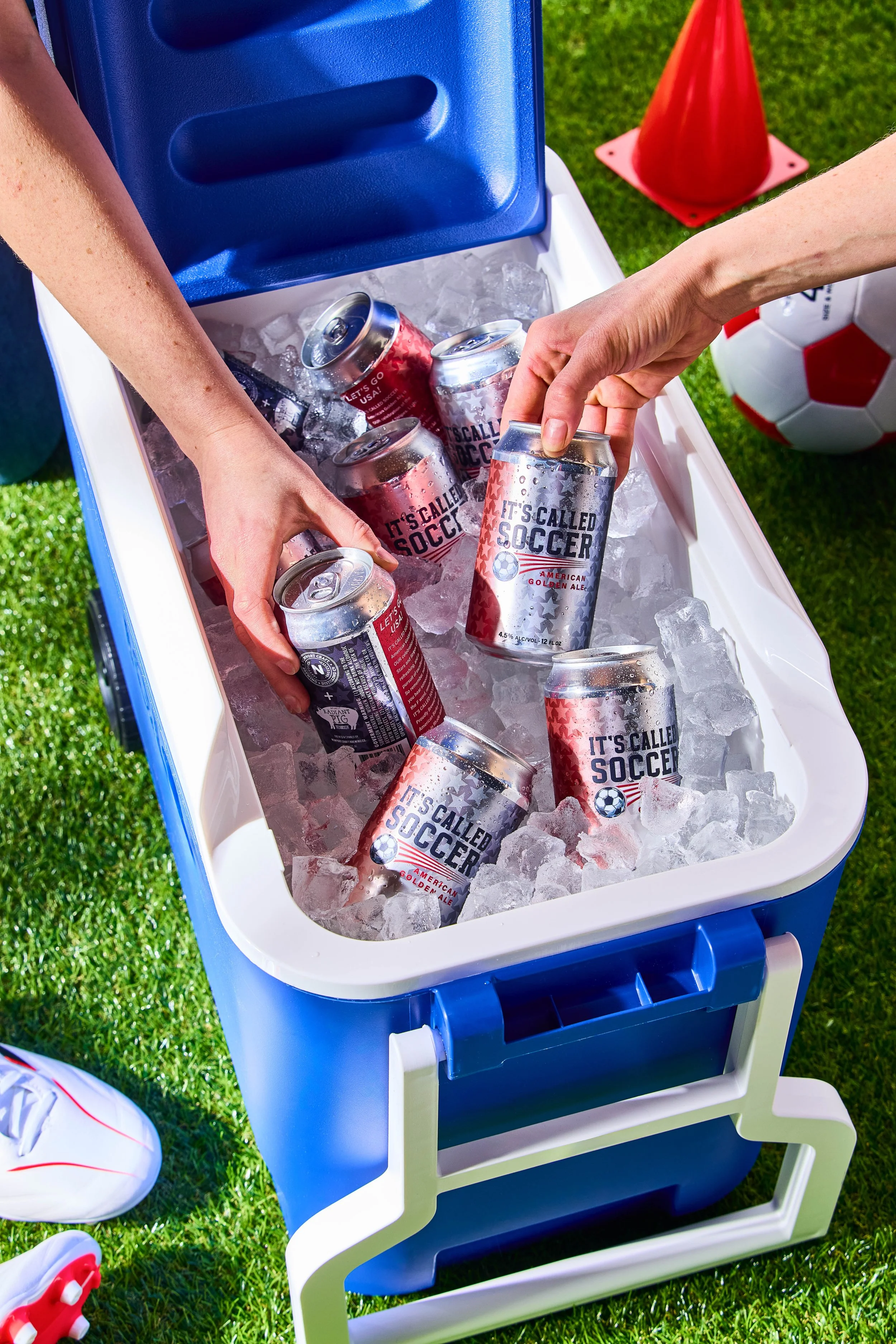 People grab cans of beer from a blue cooler filled with ice, outdoors on a grassy field near a soccer ball and orange traffic cone.