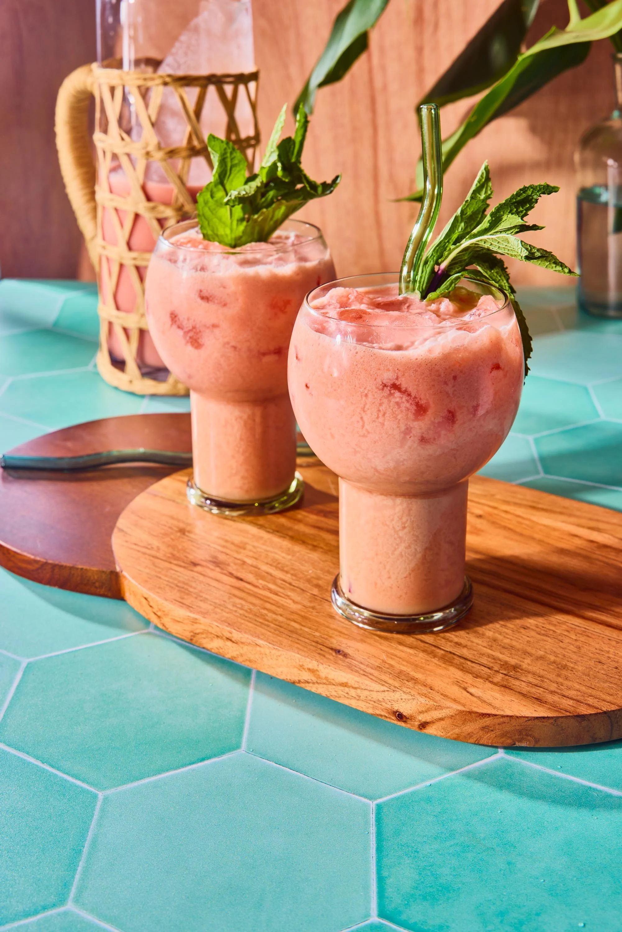 Two pink smoothies with green garnishes and glass straws on a round wooden tray, placed on a turquoise tiled surface.