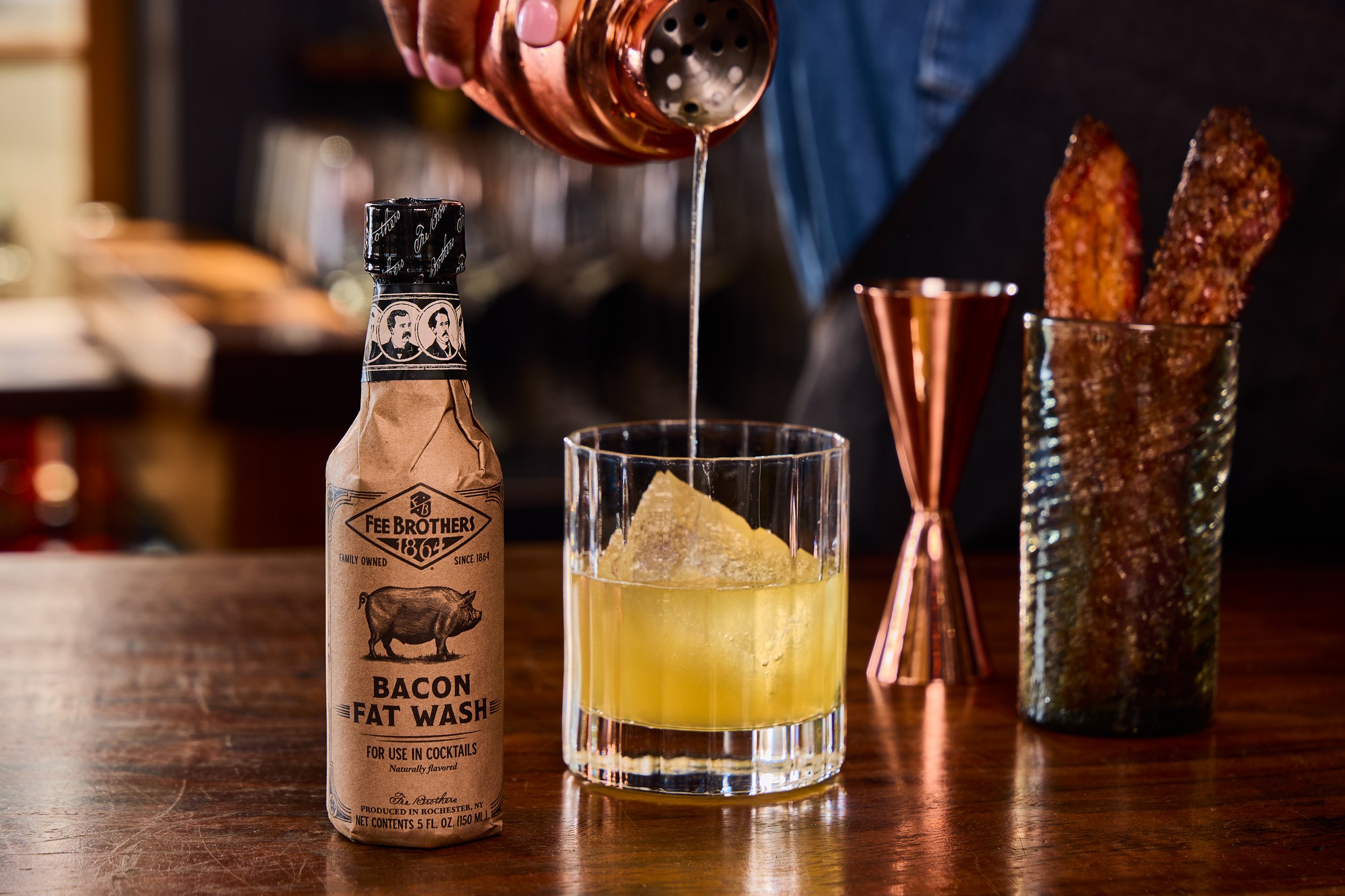 A glass of yellow cocktail with ice, a bottle of bacon fat wash, and two slices of candied bacon in a copper cup on a wooden bar or table.