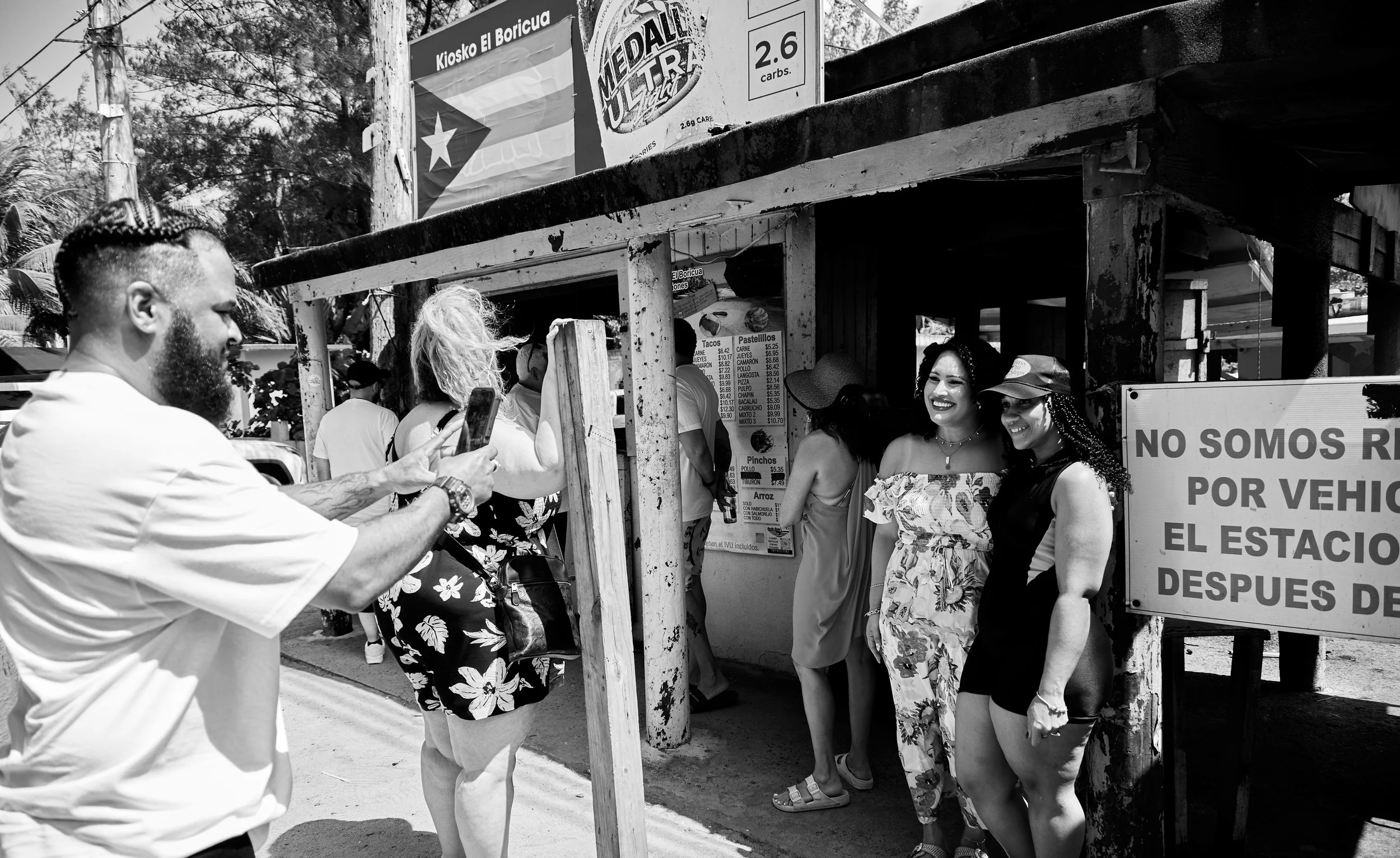 People waiting in line at a taco stand, some smiling, with a sign in Spanish and menu prices visible. One man in the foreground is taking a photo or video with his phone.