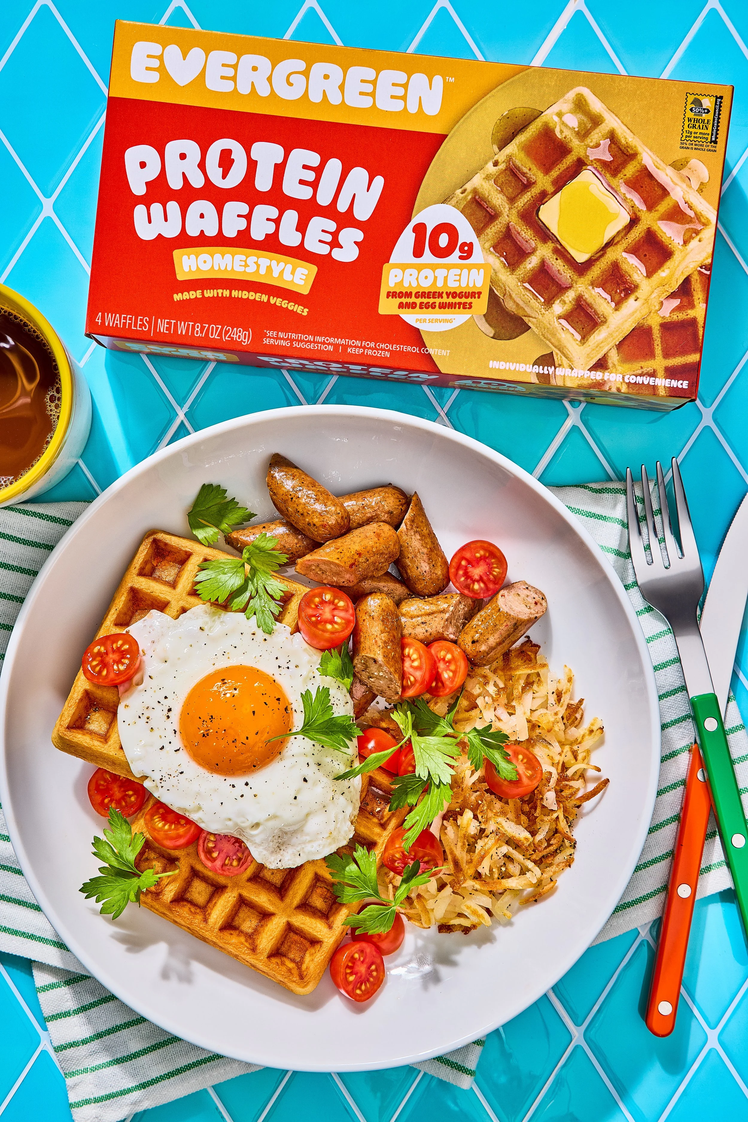 A breakfast plate with a waffle topped with a fried egg, cherry tomatoes, and parsley, along with breakfast sausage links and hash browns, on a white plate. There is a box of Evergreen Protein Waffles, a yellow mug, and a fork and knife on a green-st