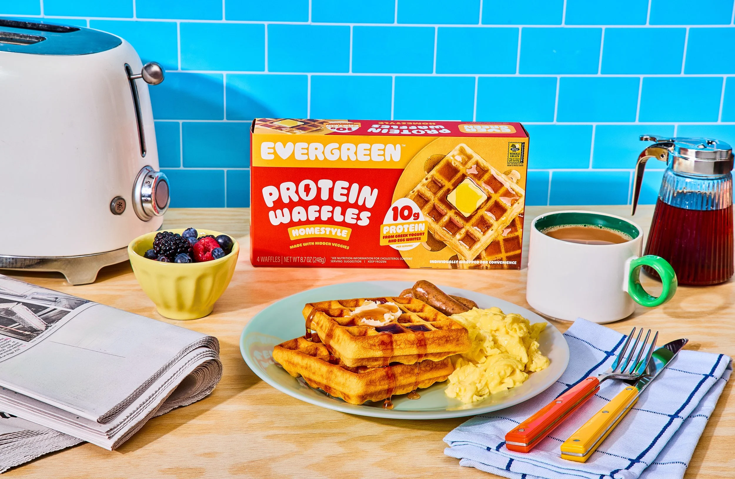 Breakfast table with waffles, scrambled eggs, sausage, a bowl of mixed berries, a mug of coffee, a syrup dispenser, a box of protein waffles, an open newspaper, and colorful utensils on a blue and white striped napkin.