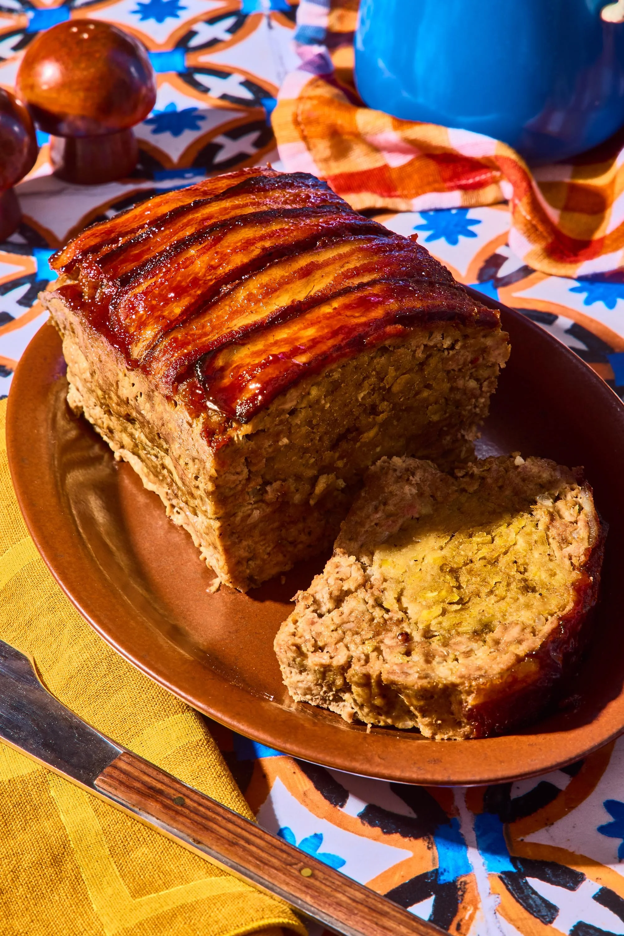 A freshly baked meatloaf glazed with ketchup sits on a brown plate, with a slice cut revealing its mixture of ground meat and vegetables. The plate is on a colorful tablecloth with a vibrant pattern, accompanied by a yellow napkin and a wooden-handle