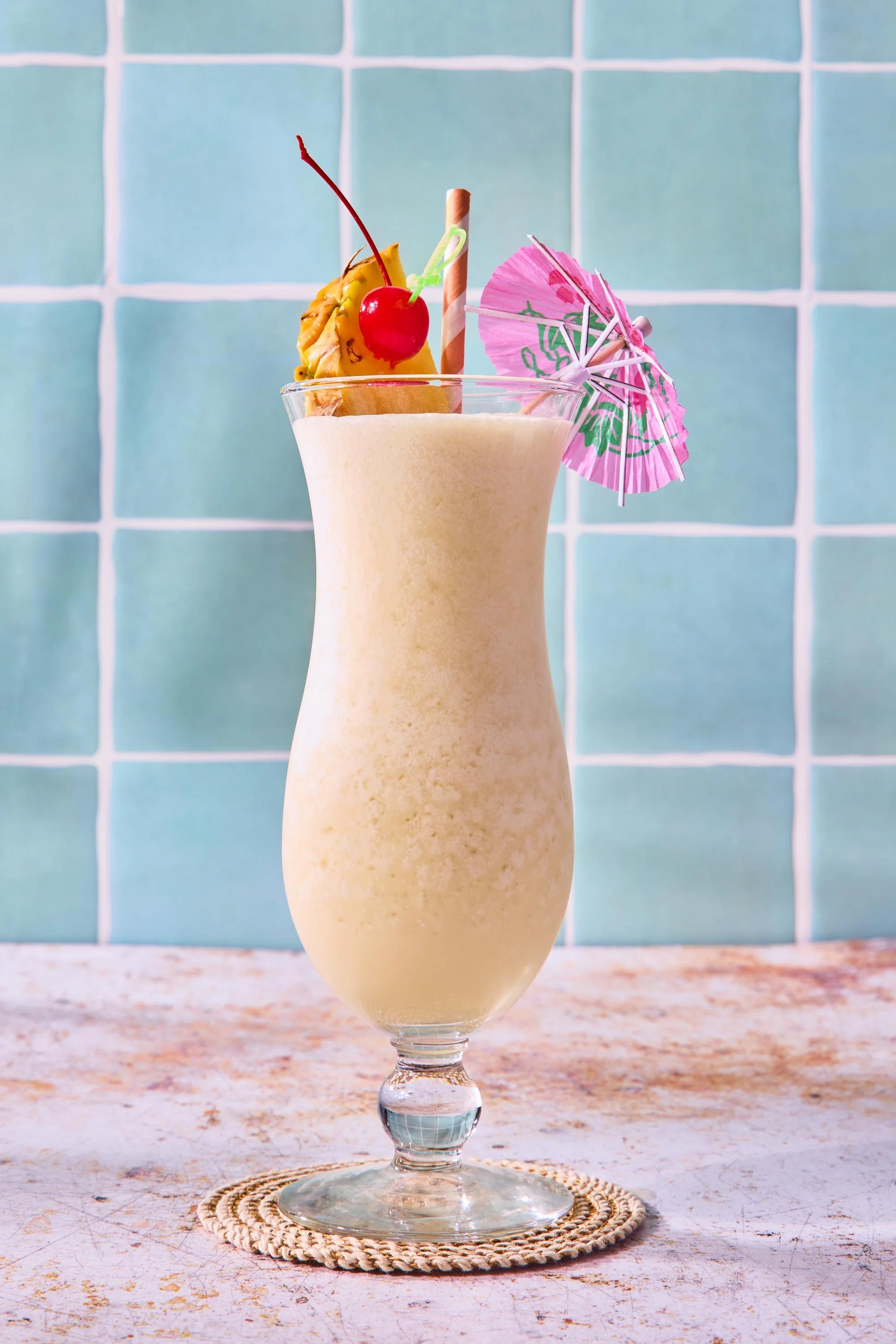 A tall glass of a frothy, creamy beverage garnished with a pineapple chunk topped with a maraschino cherry, a small decorative pink paper umbrella, and a striped straw. The glass sits on a woven coaster on a rustic surface against a background of blu