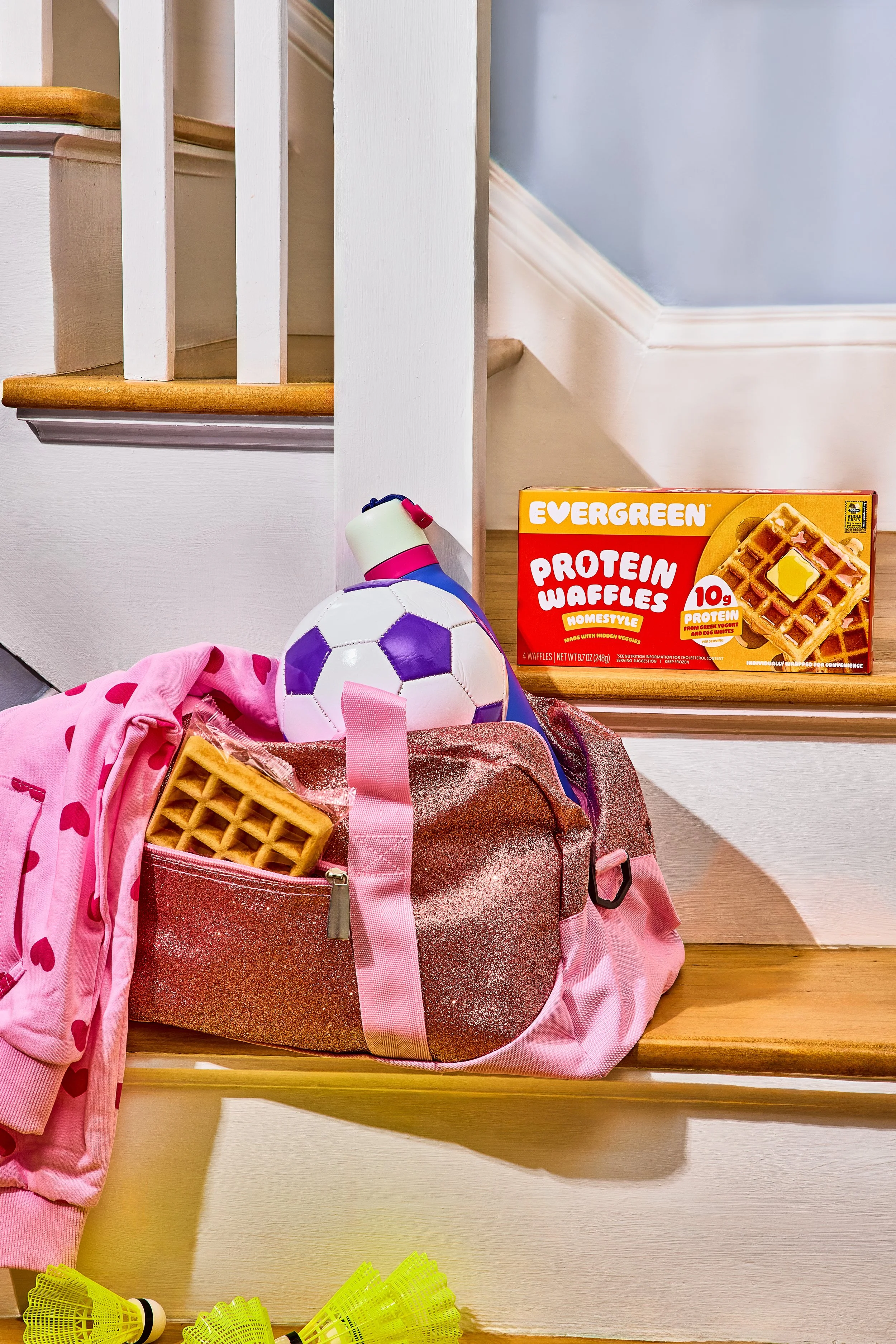 A pink backpack with sparkles, a pink bag with hearts, a soccer ball, a waffle, and a box of protein waffles on stairs. There are yellow badminton birdies on the floor.